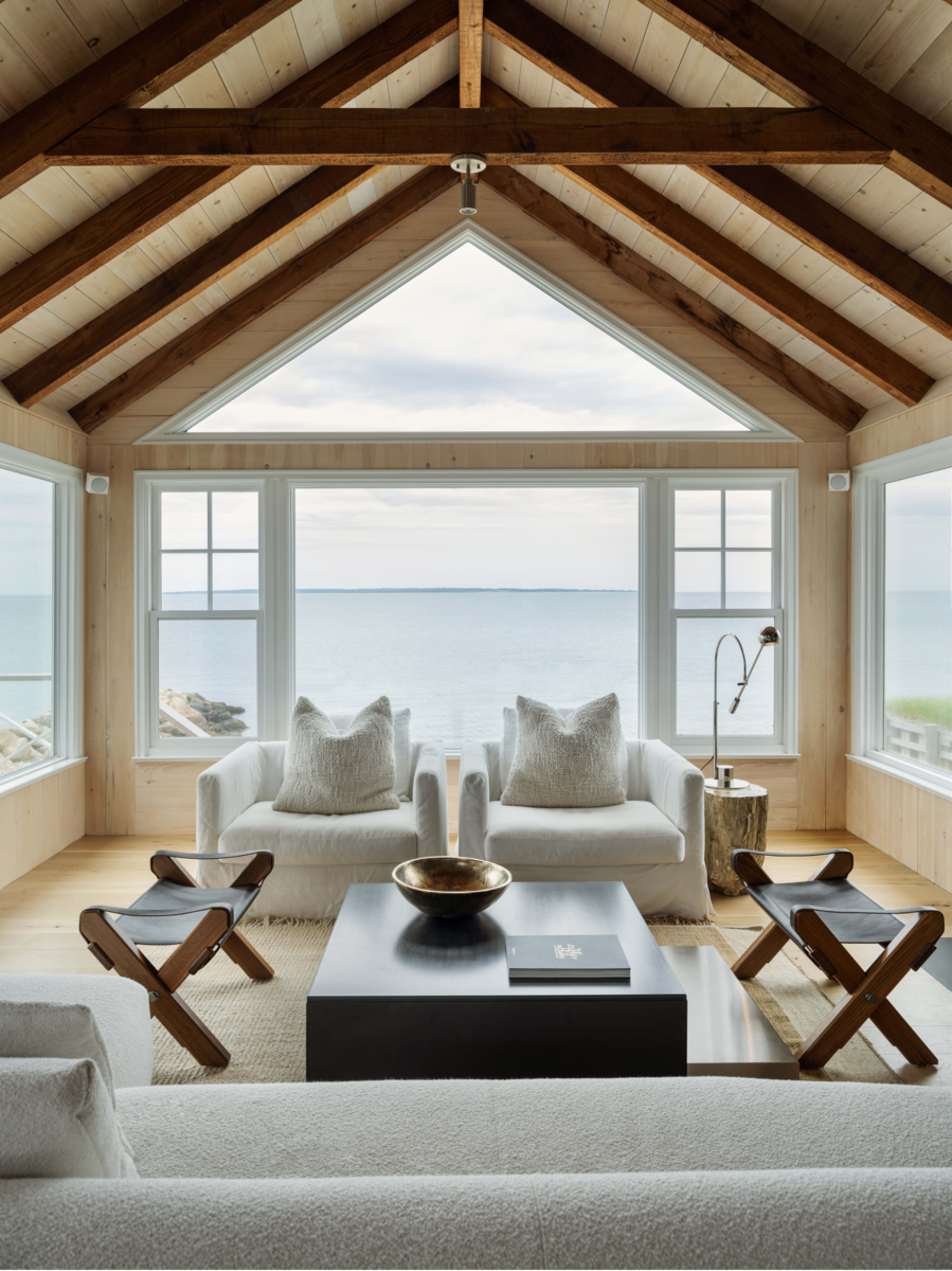 A bright living area features large windows overlooking a calm body of water, with minimalist furniture arranged around a central table.