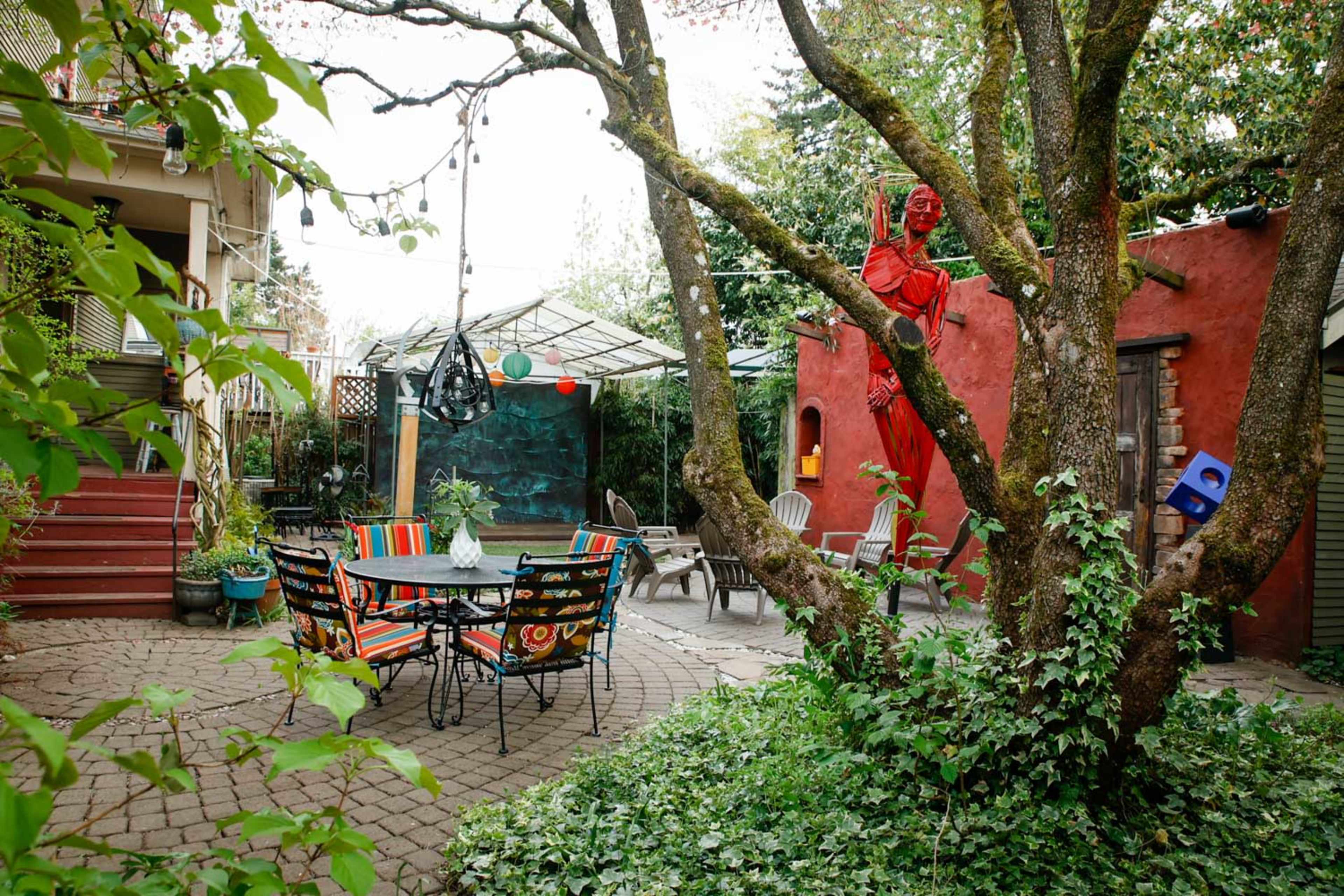 A courtyard features a circular table surrounded by colorful chairs, with a hanging swing and a painted wall in the background.