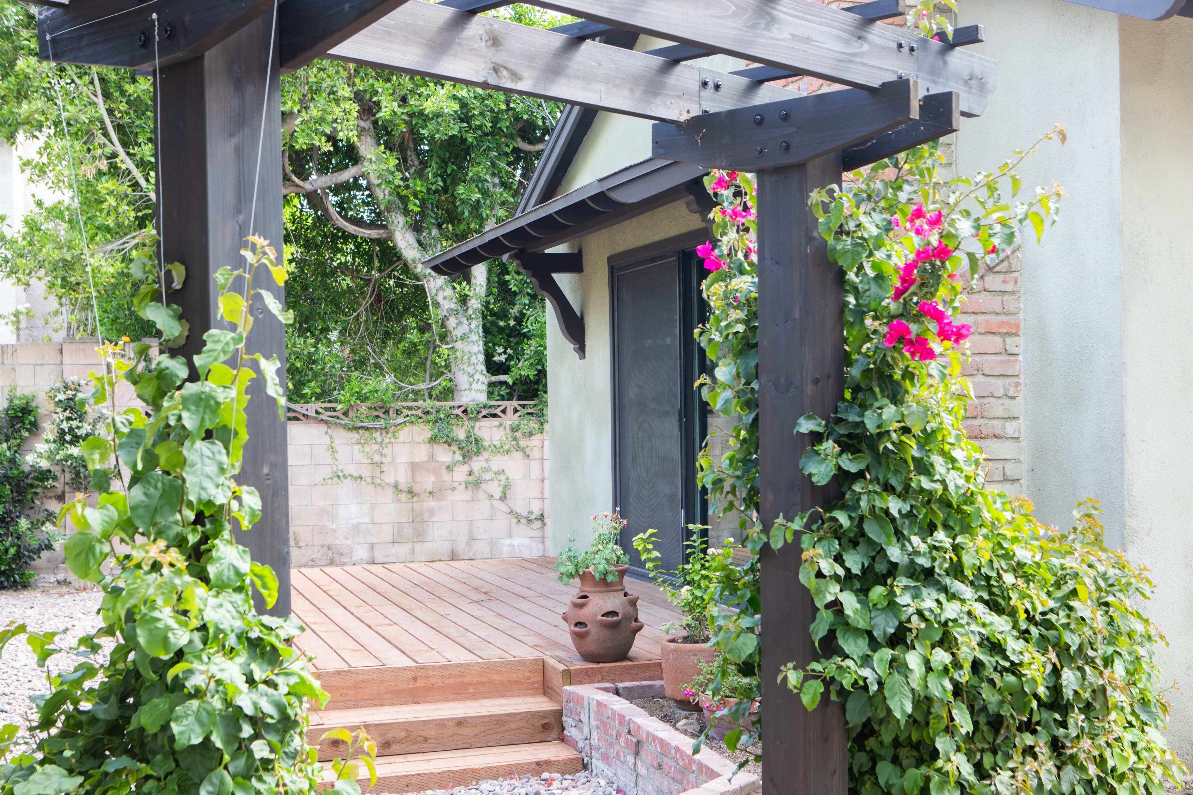 The image shows a wooden pergola with climbing plants and flowers leading to a small wooden deck and a light-colored house in the background.