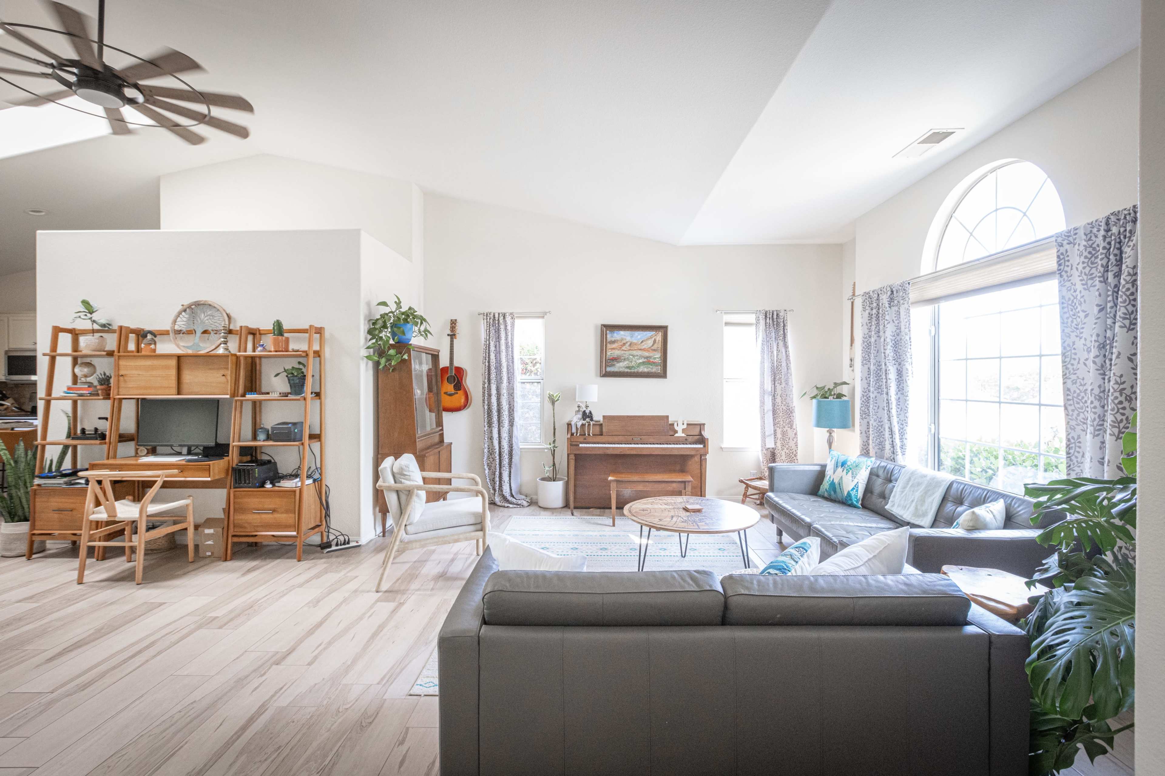 A bright living room features a seating area with a gray sofa, a wooden coffee table, a desk, and a piano, with large windows allowing natural light to enter.