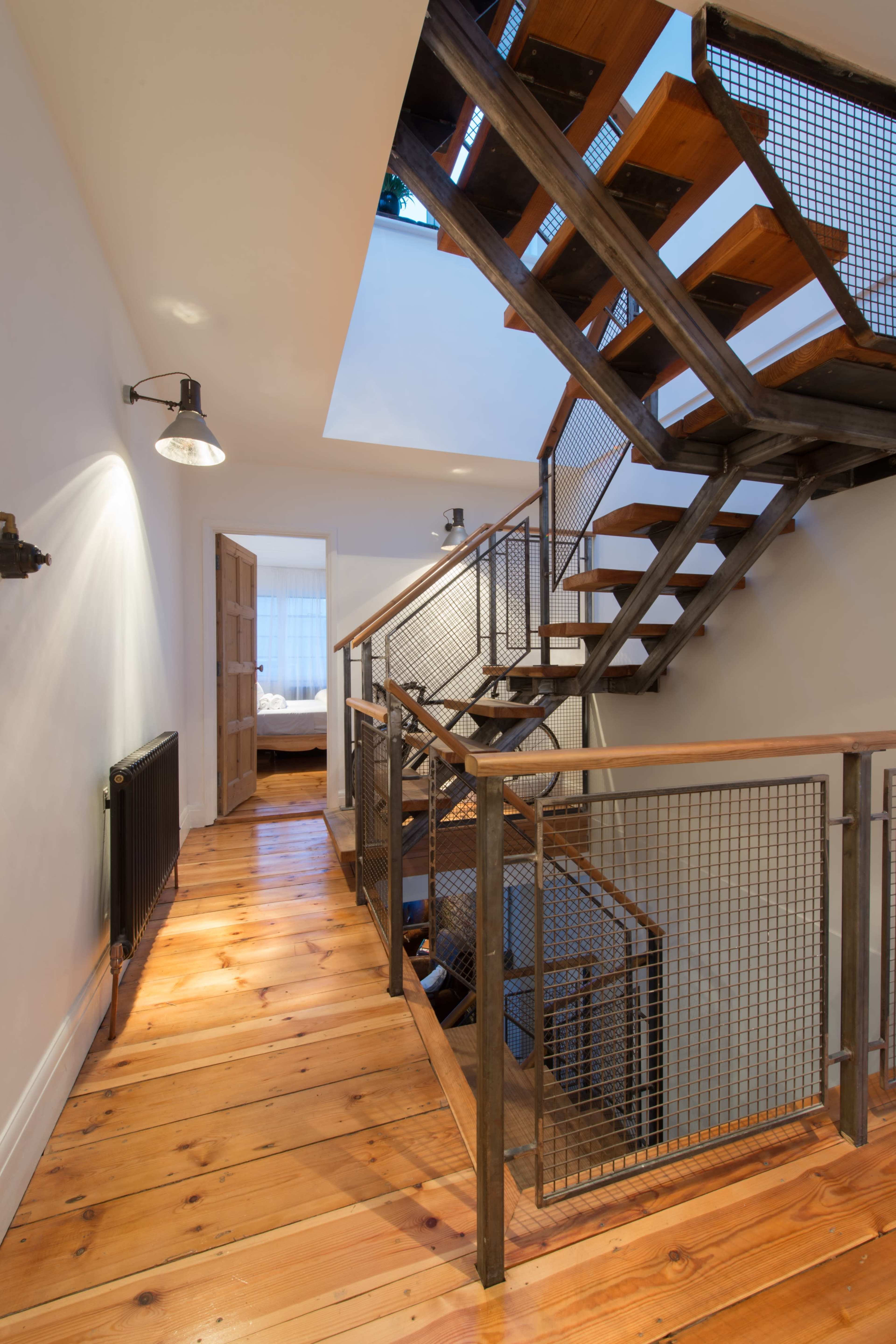 The image shows a modern staircase made of wood and metal, with a wooden floor and a hallway leading to a door and a room in the background.