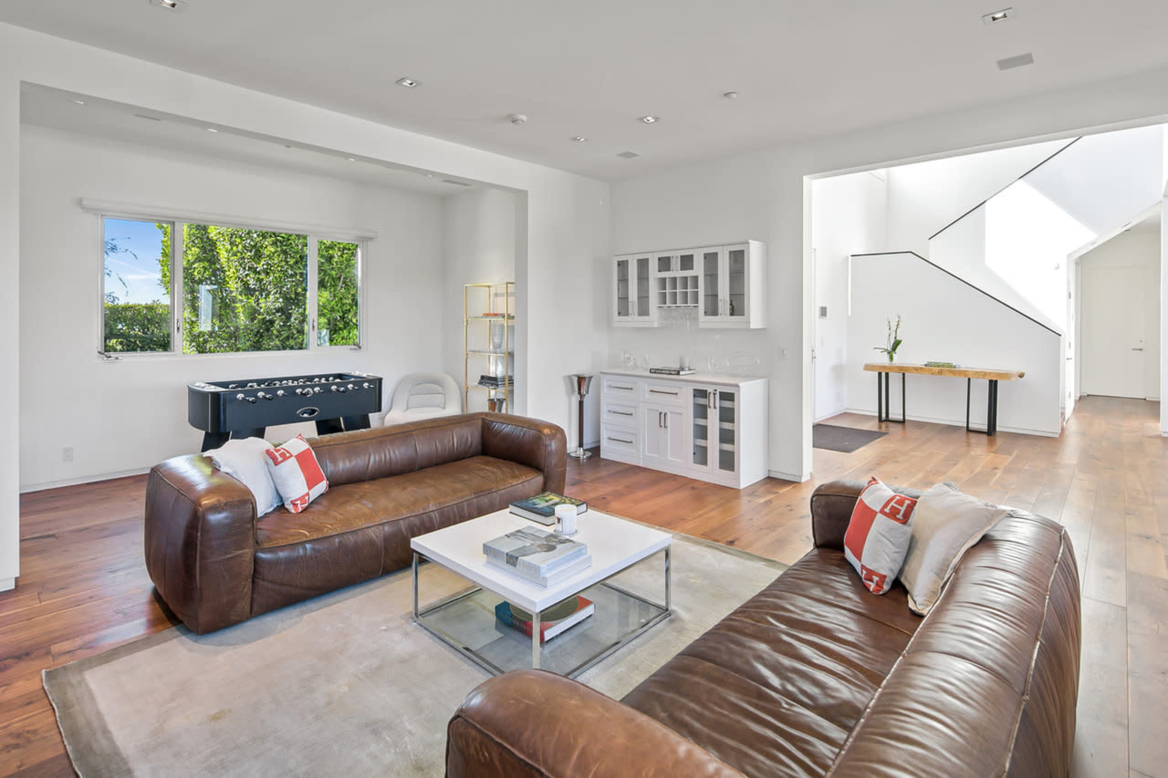 The image shows a modern living room with two brown leather sofas, a glass coffee table, and a kitchenette in the background, featuring large windows that allow natural light to enter the space.