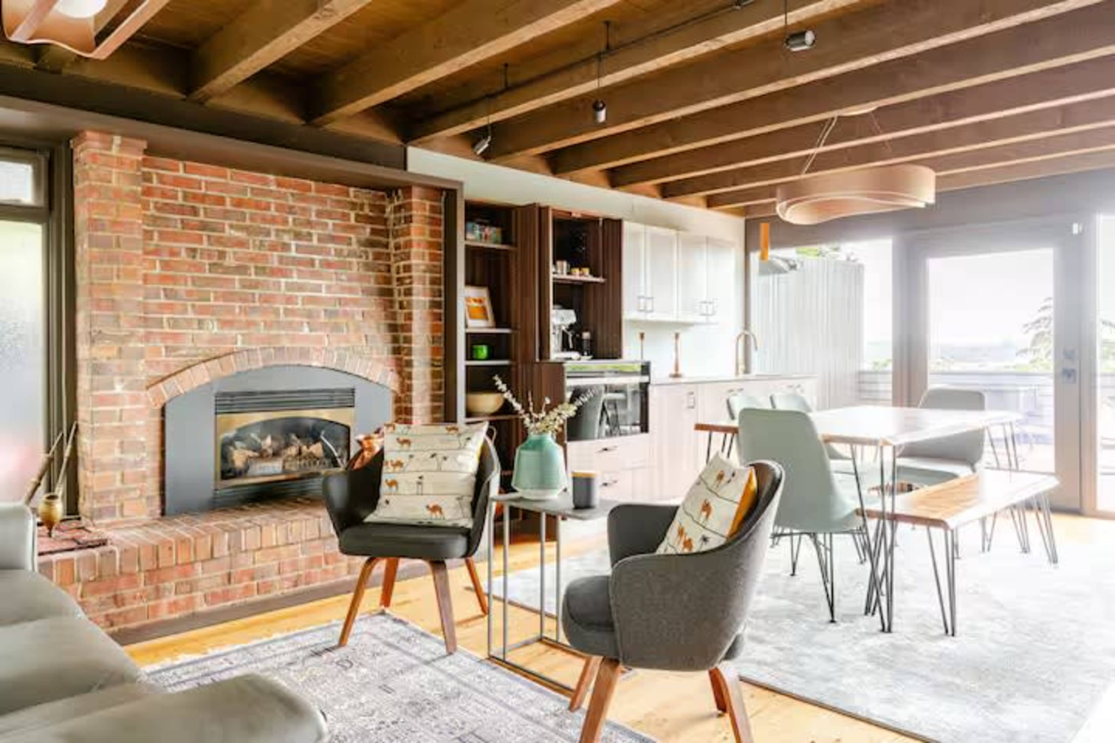 The image showcases a cozy living area featuring a brick fireplace, modern furniture, and a dining space with large windows that open to an outdoor area.