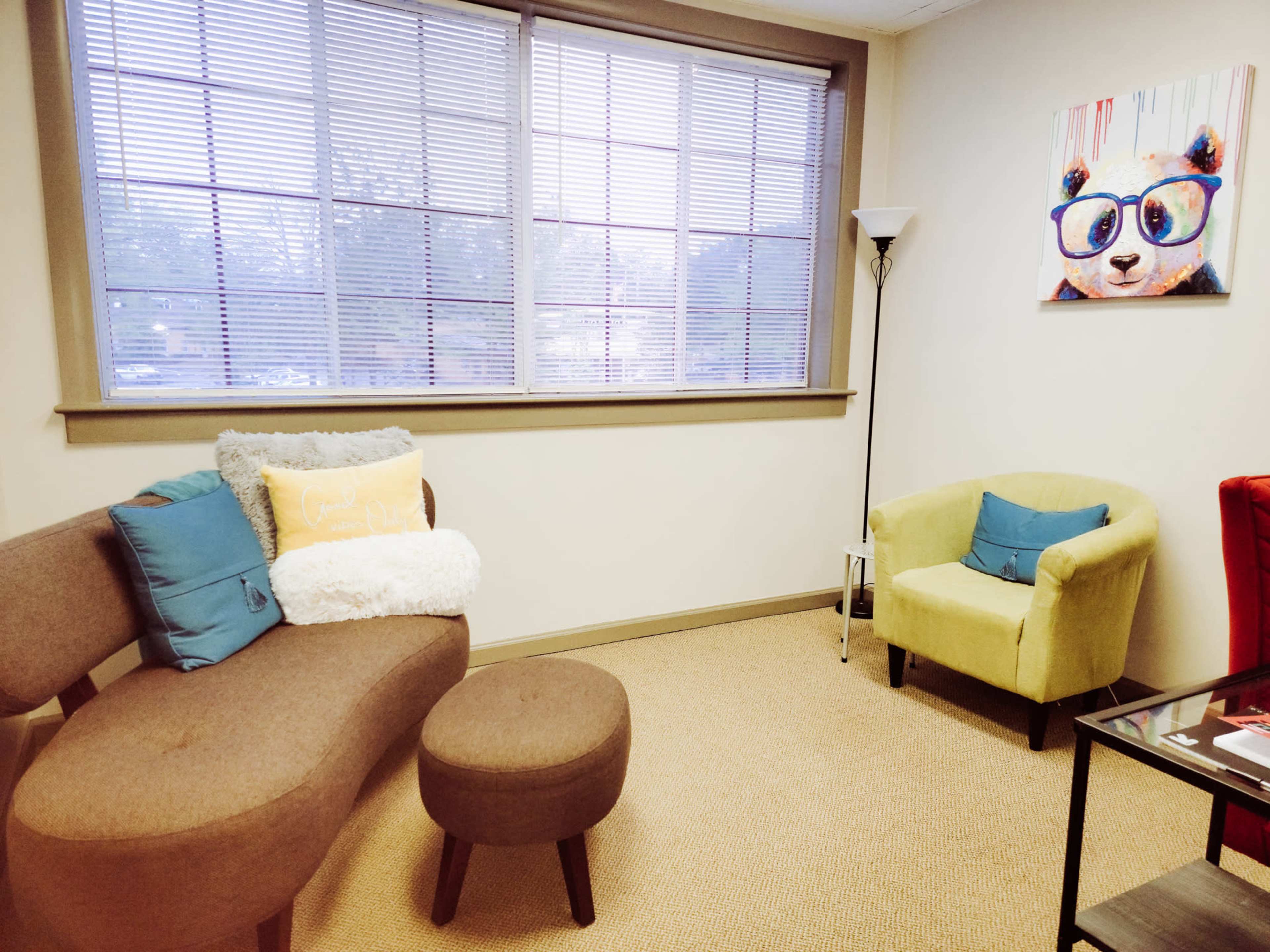A cozy office space with a curved sofa, an ottoman, and two armchairs, featuring a lamp and a colorful panda painting on the wall.