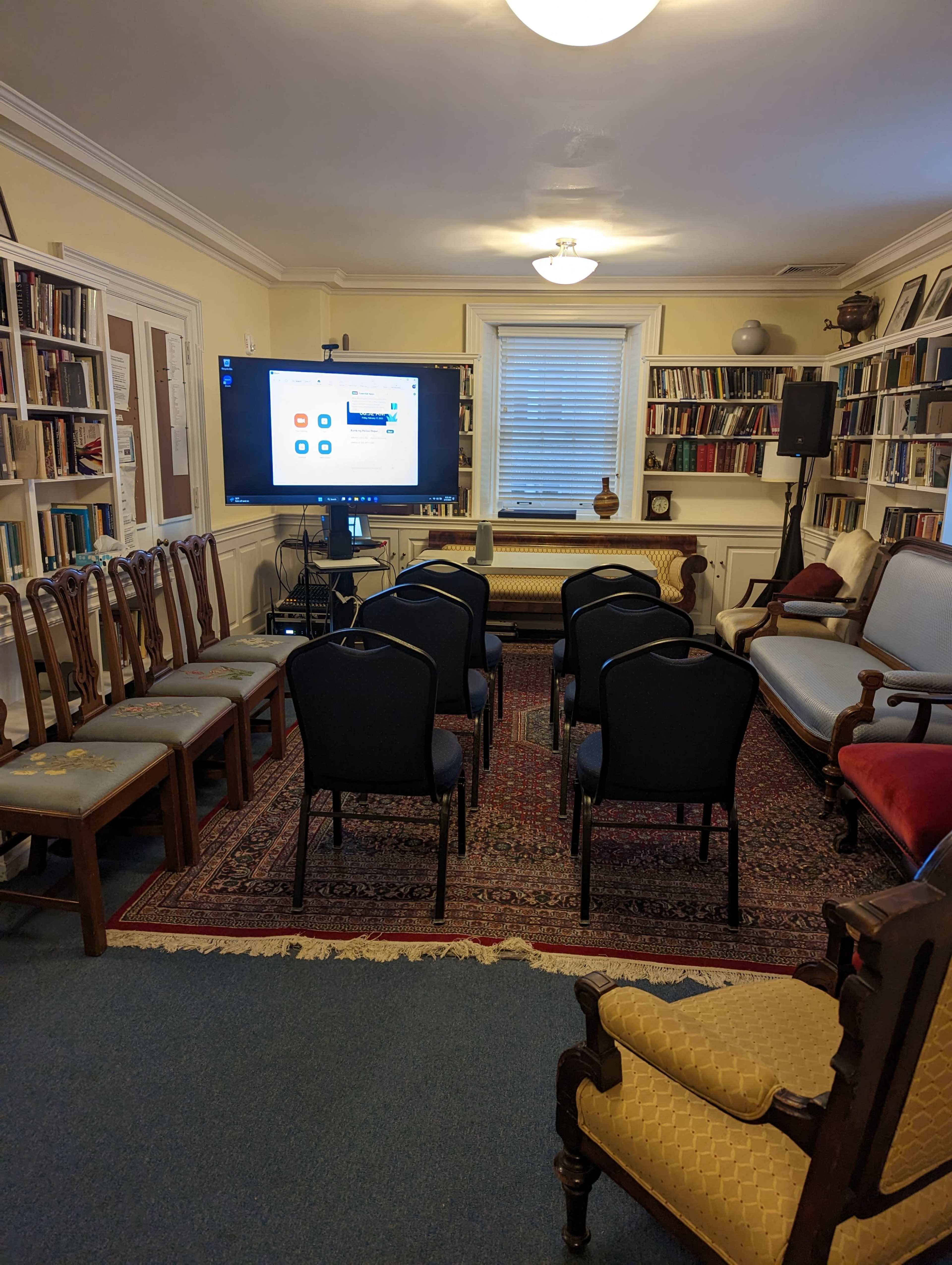 A small room filled with bookshelves contains several rows of chairs facing a large screen displaying a video call interface.