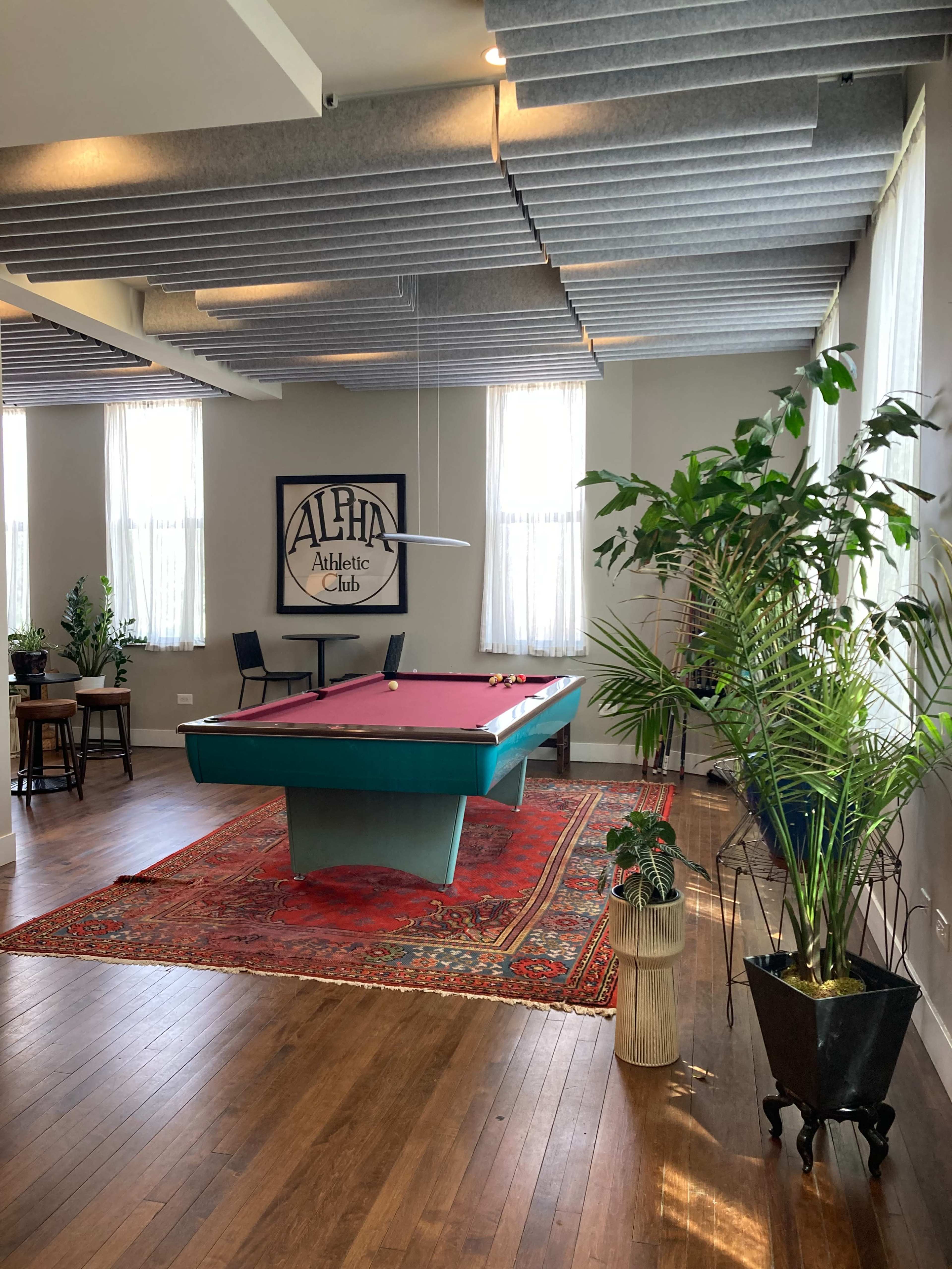 The image shows a spacious room with a red pool table, a large area rug, plants, and a sign that reads "Alpha Athletic Club" on the wall.