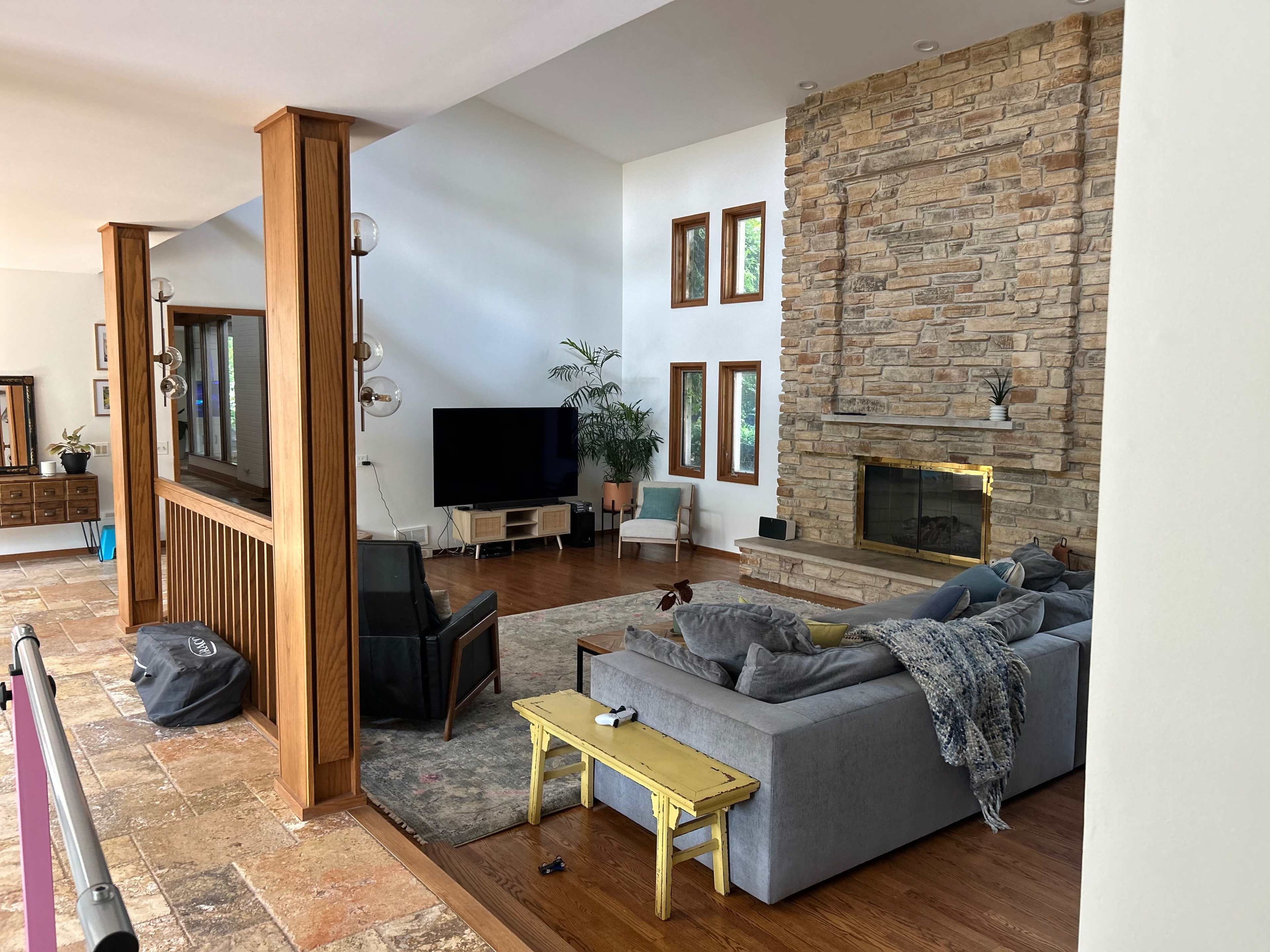 A modern living room with a stone fireplace, large windows, a gray sofa, and a yellow coffee table.
