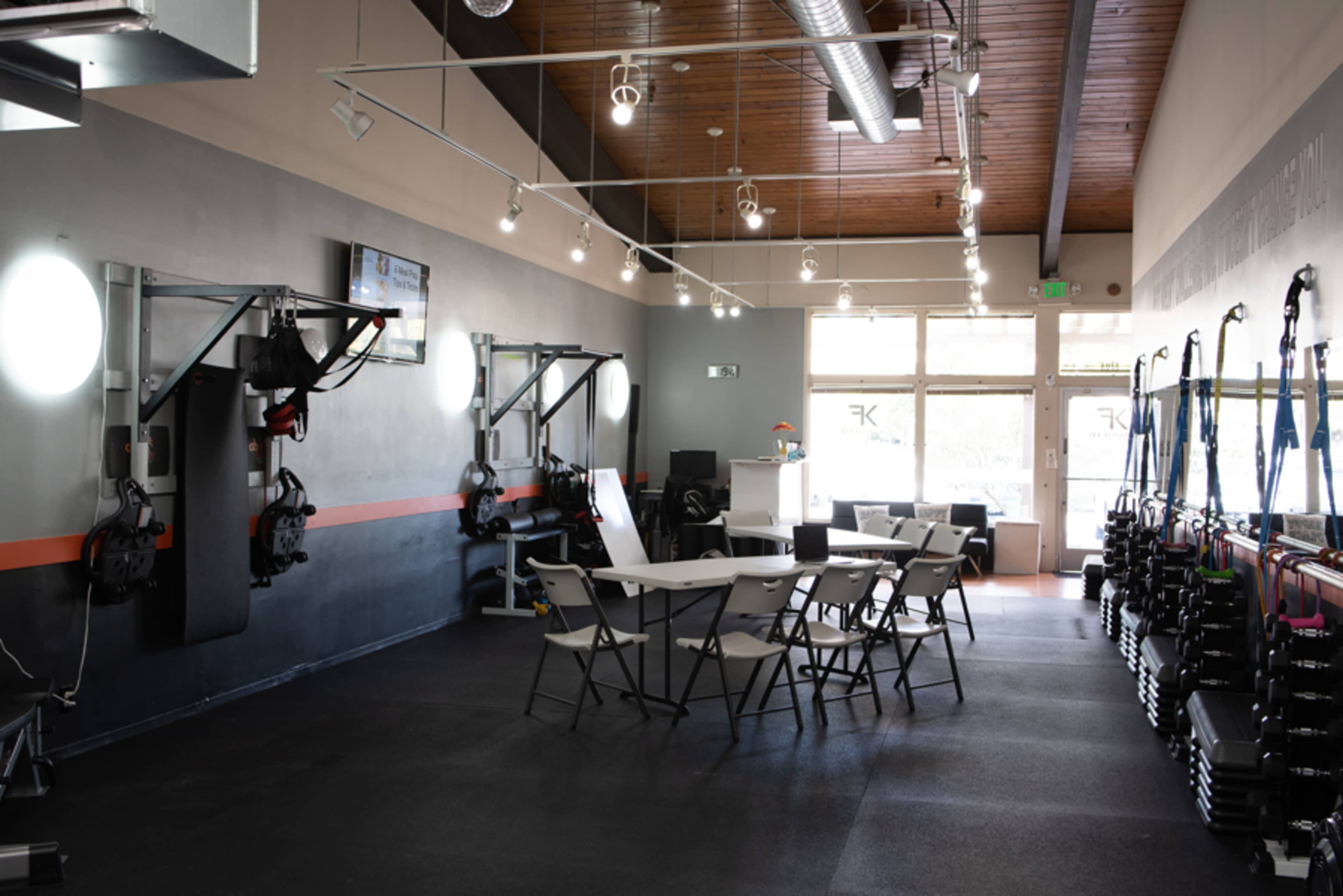 The image shows a modern gym interior with exercise equipment on one side and a meeting area with a table and chairs on the other.