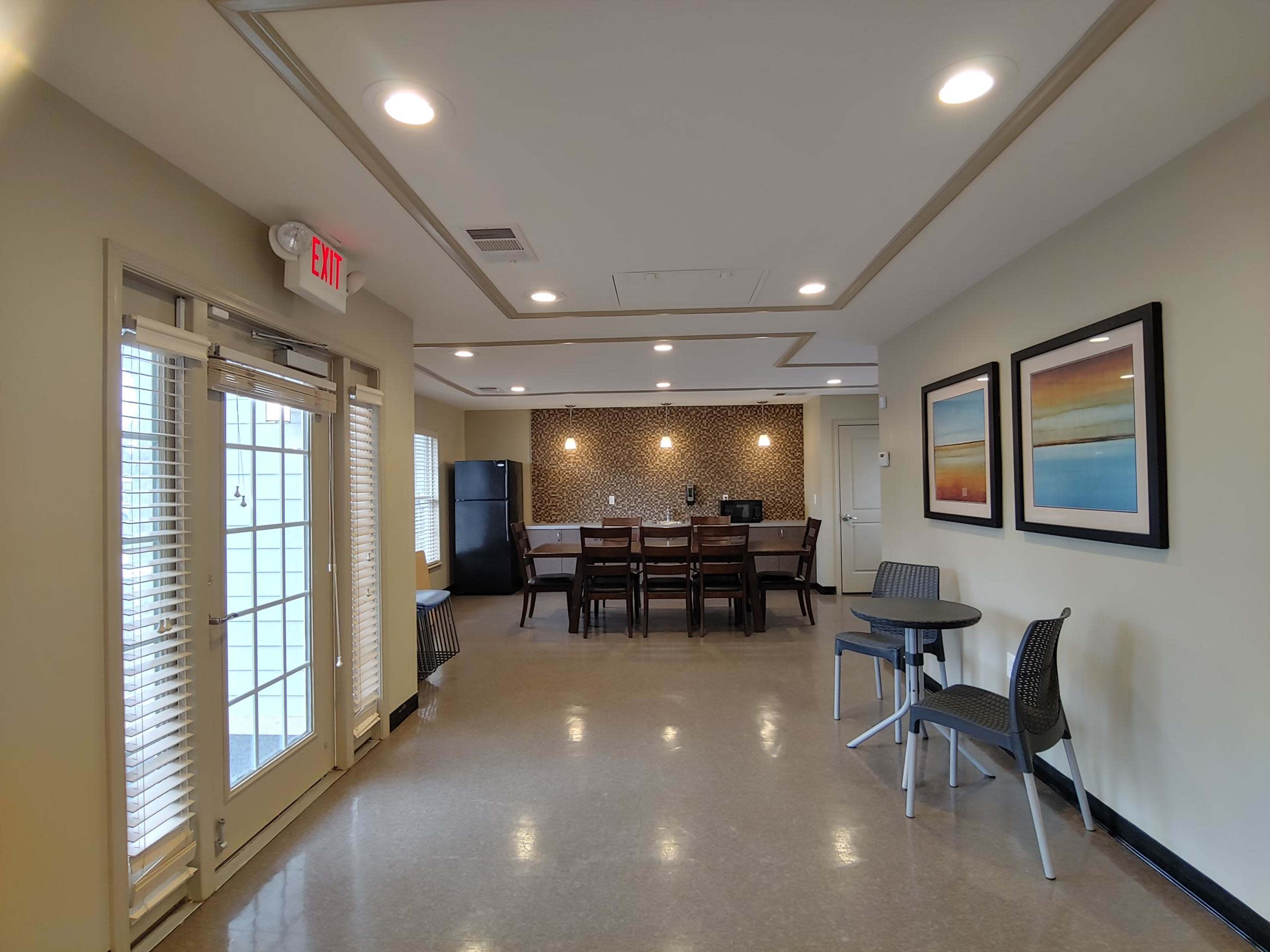 The image shows a modern communal kitchen area featuring a dining table, chairs, and a kitchenette with a refrigerator, as well as access to an outdoor patio through double doors.