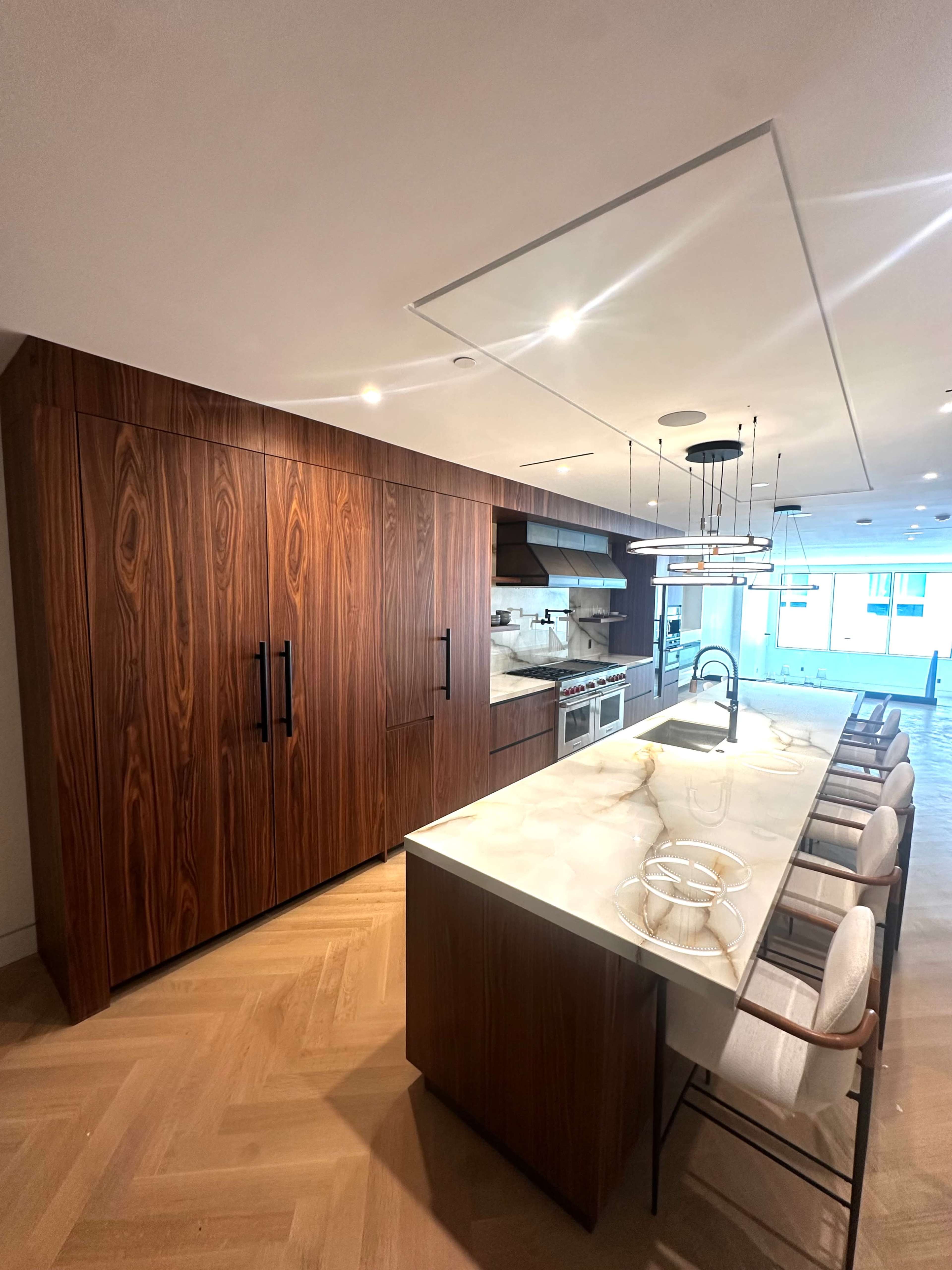 The image shows a modern kitchen featuring dark wooden cabinetry, a large marble island with seating, and stainless steel appliances.