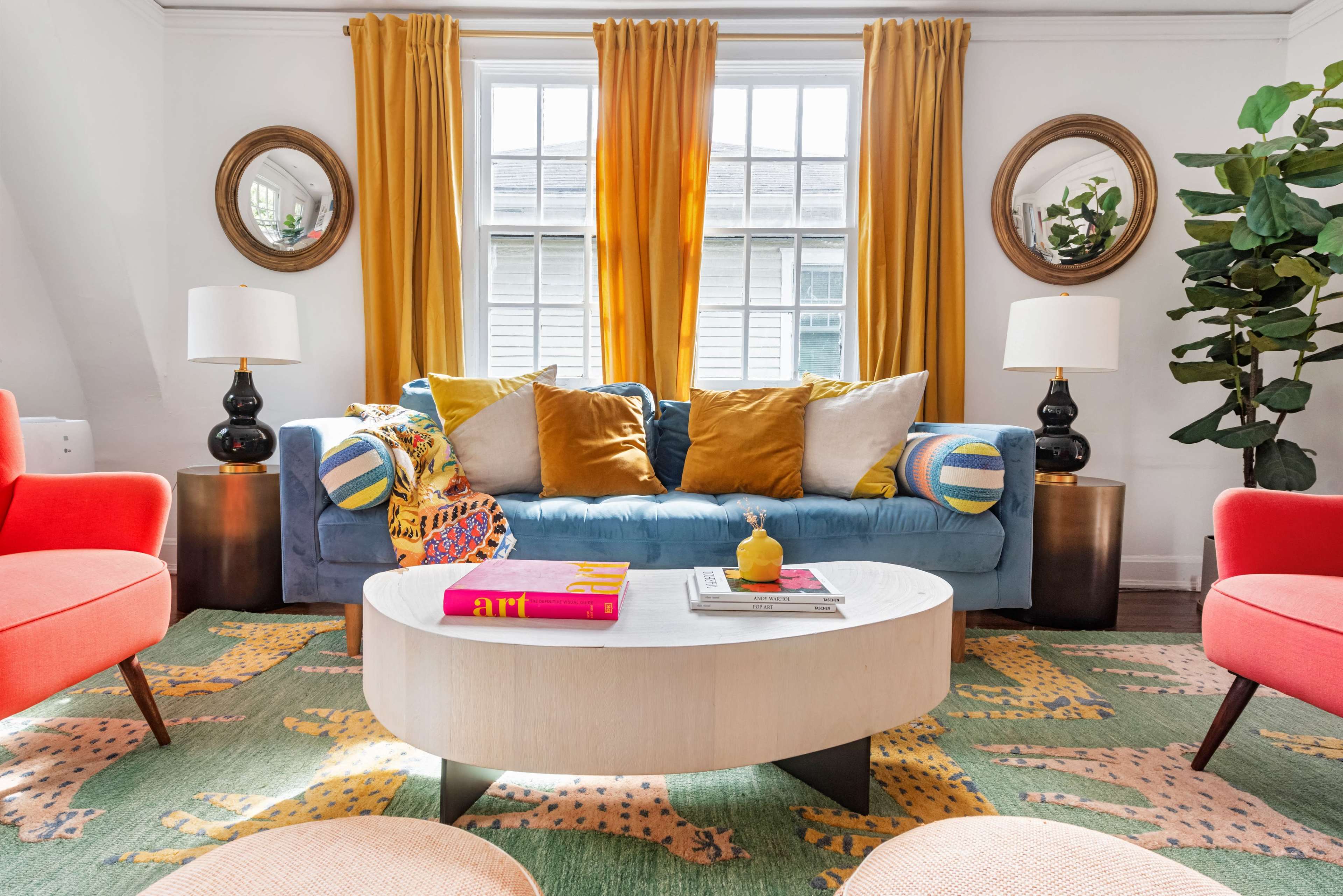 The image features a brightly decorated living room with a blue sofa, colorful cushions, a round coffee table, and two red armchairs, all set against a patterned rug and large windows.