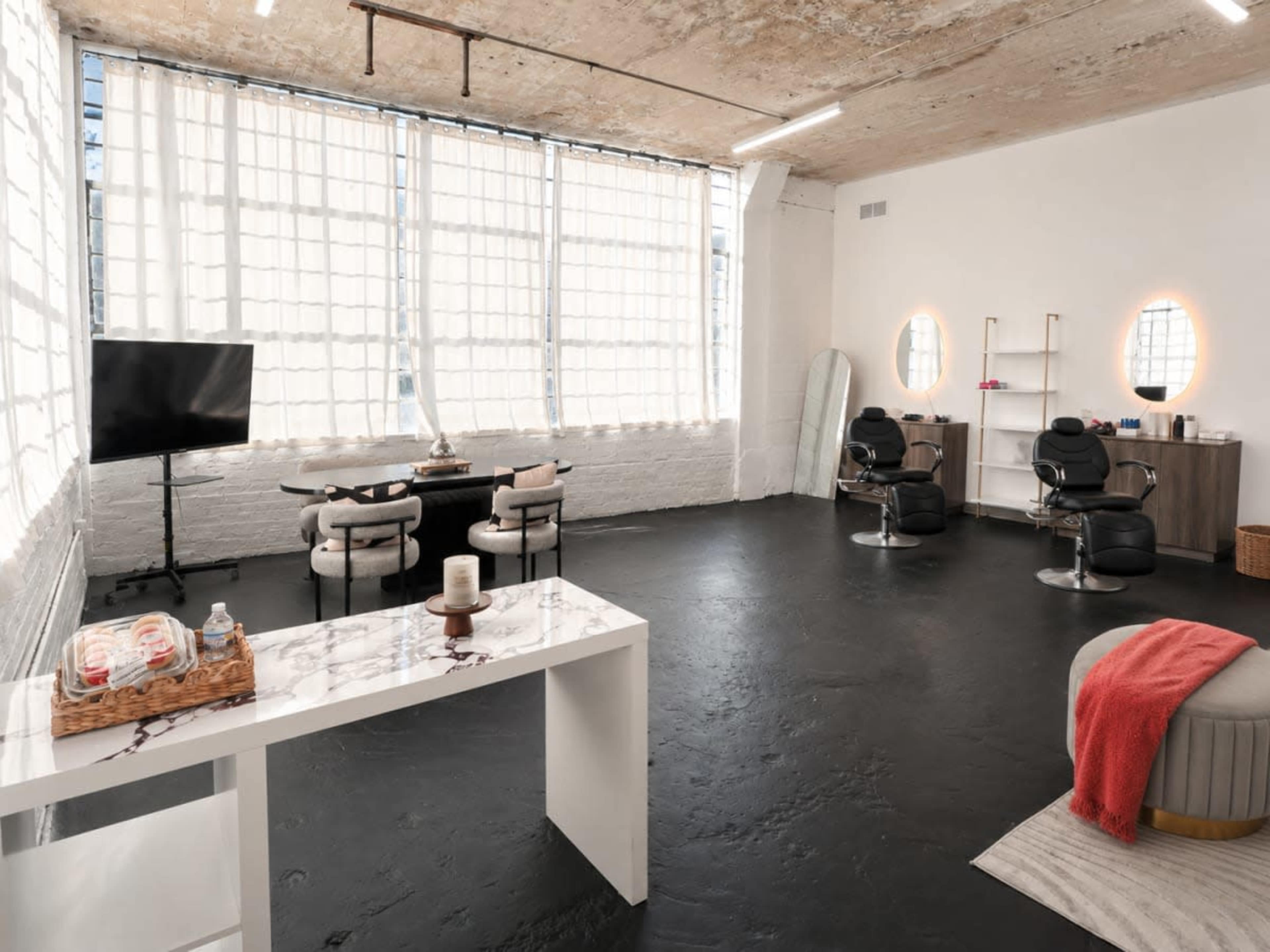 The image depicts a spacious, well-lit hair salon featuring two styling chairs, a table, and a large window with natural light.