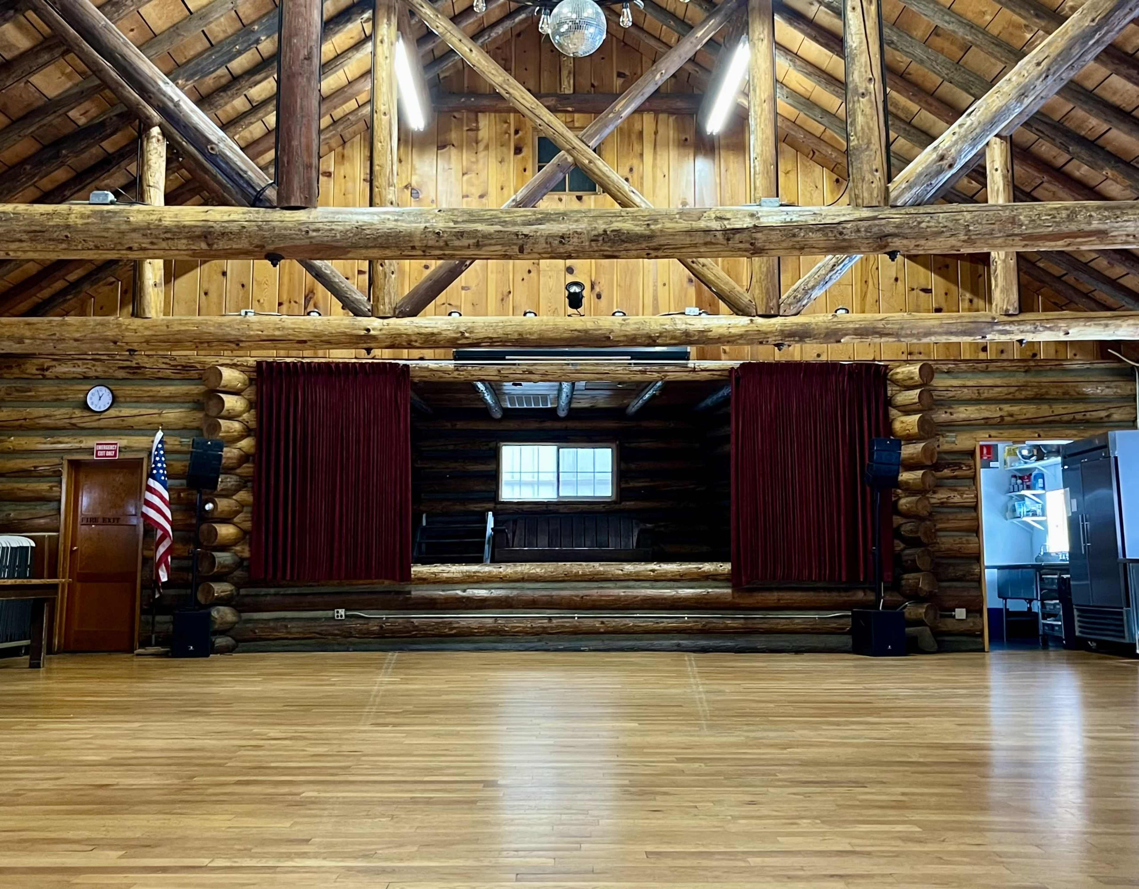The image depicts a spacious wooden hall with a high, exposed beam ceiling, a stage at the front draped with red curtains, and an American flag displayed to the left.