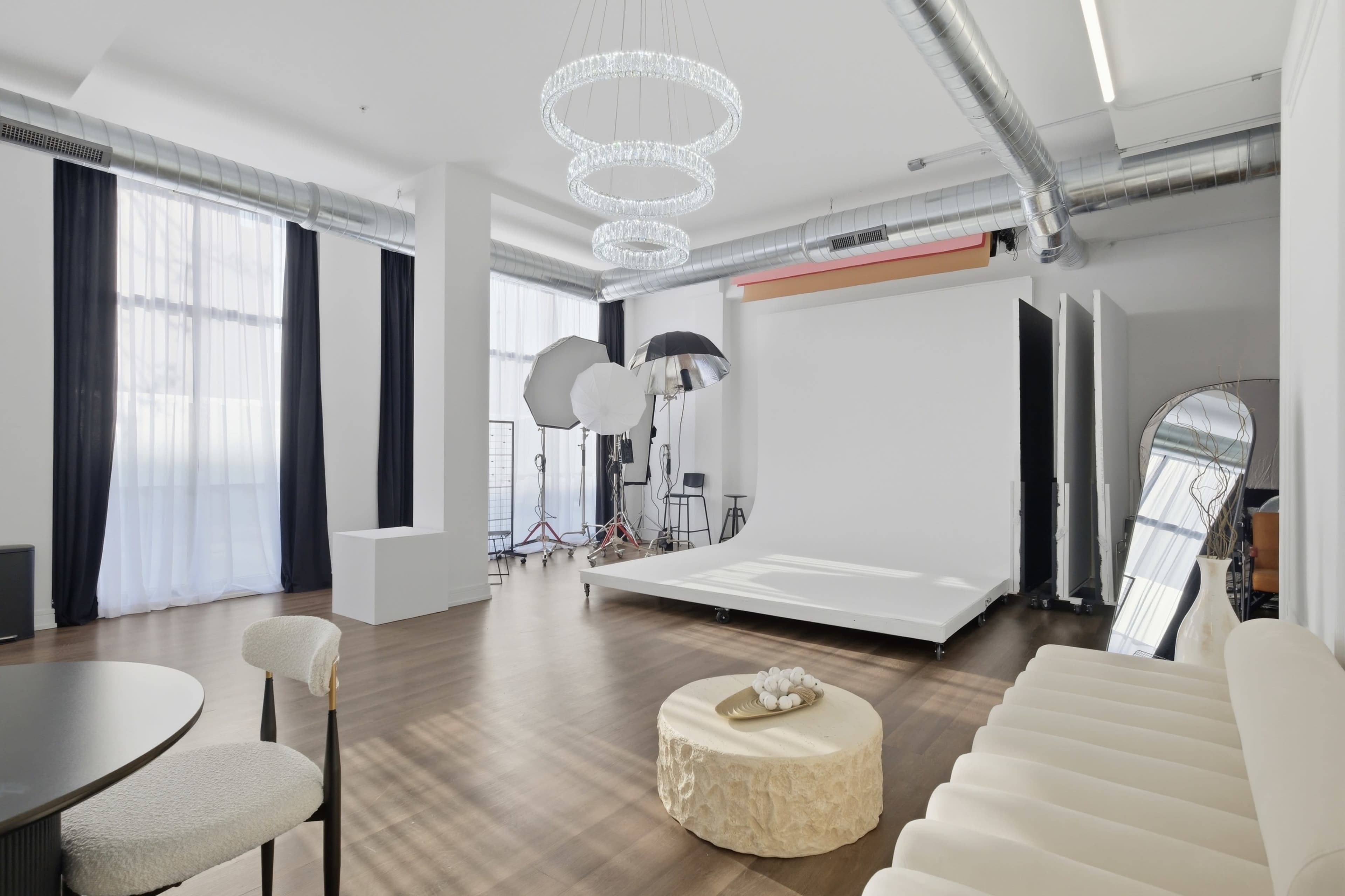 The image shows a well-lit photography studio featuring a large white backdrop, lighting equipment, and modern furnishings.
