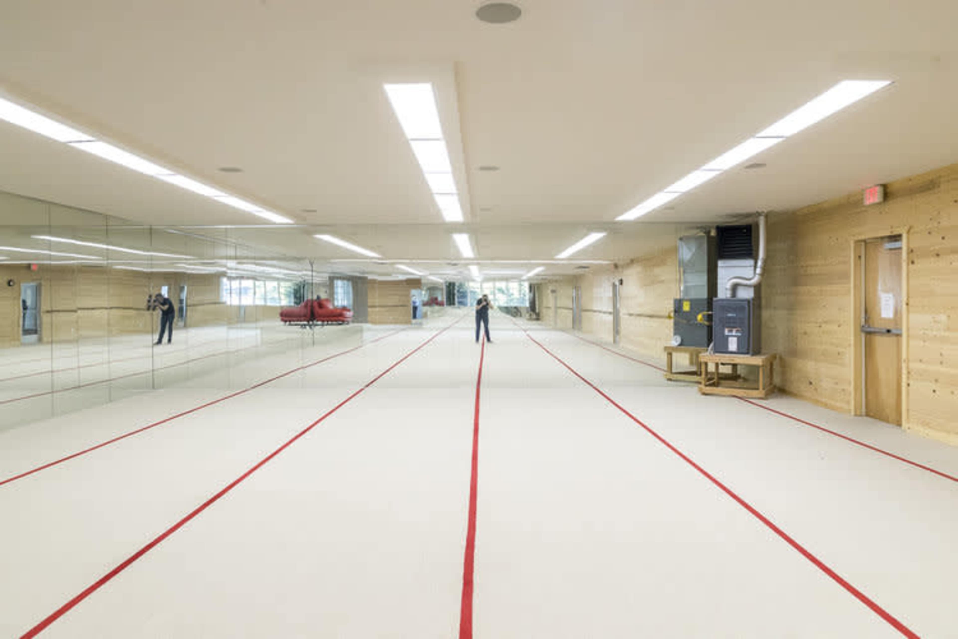 A spacious gymnasium features a mirrored wall, a red-striped floor, and ample lighting, with a person standing at the far end.