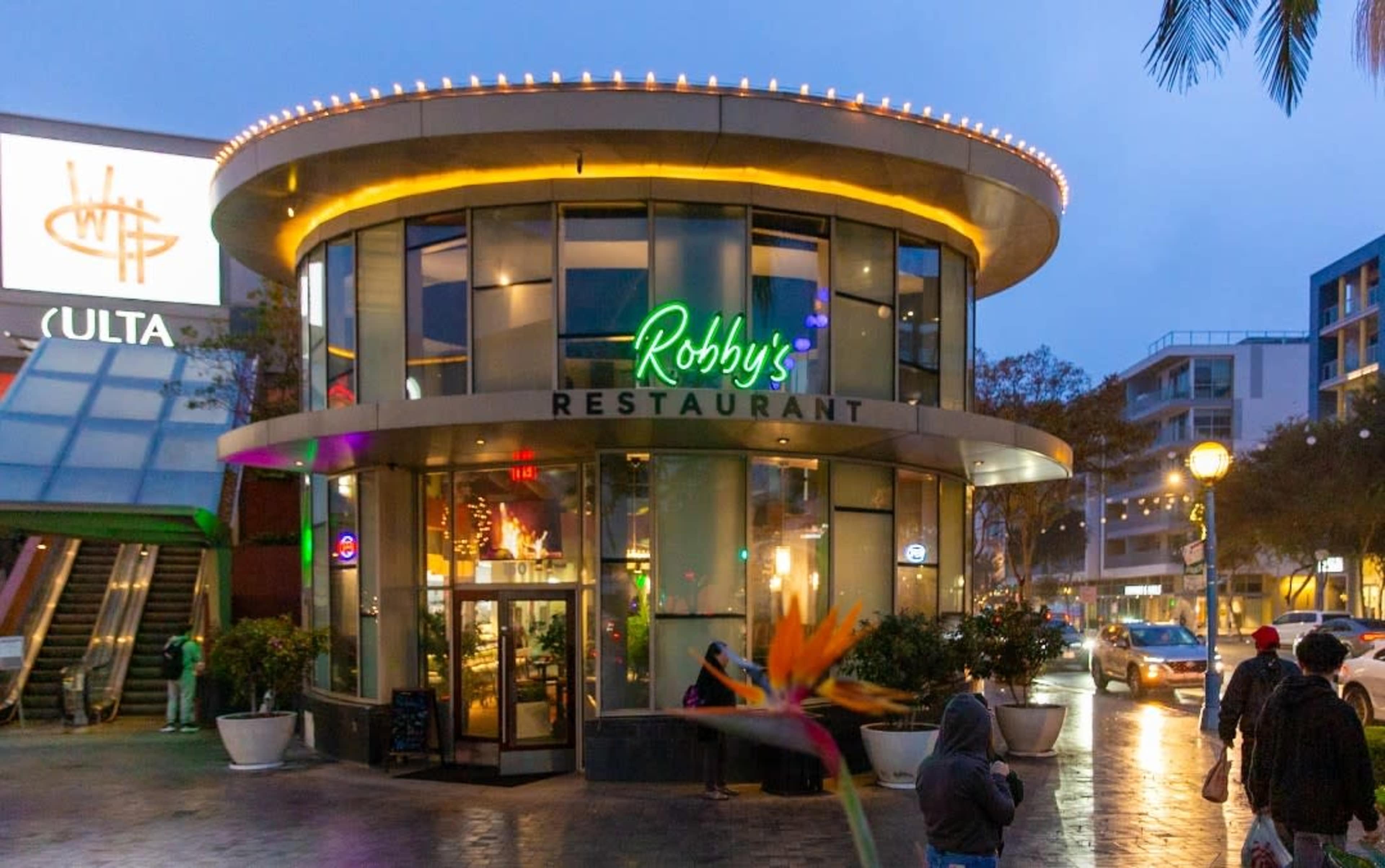 The image shows a modern circular restaurant with large glass windows, brightly lit signage, and people walking in a shopping area on a rainy evening.