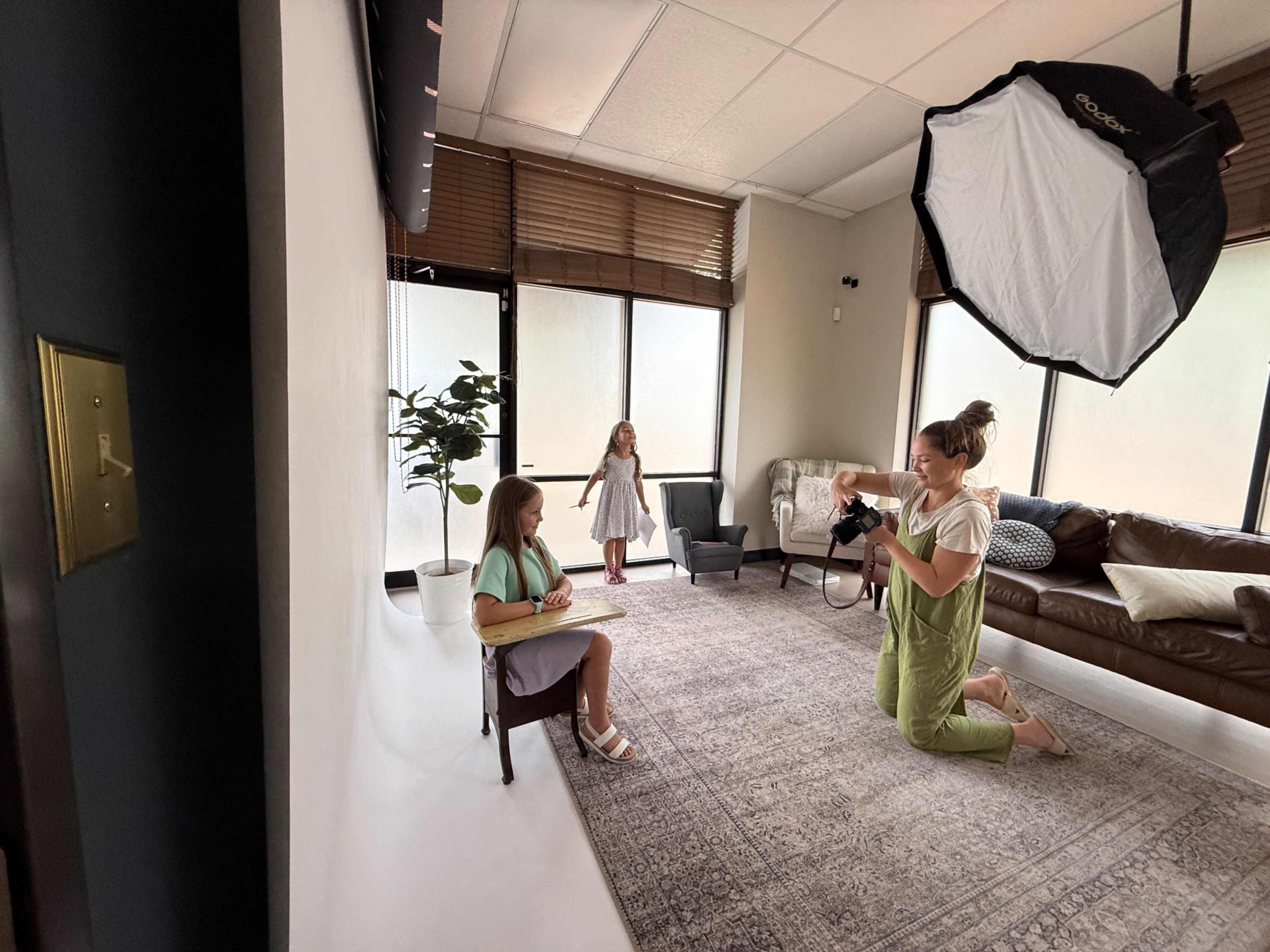 A photographer is capturing images of two girls in a well-lit studio with a sofa and a plant in the background.