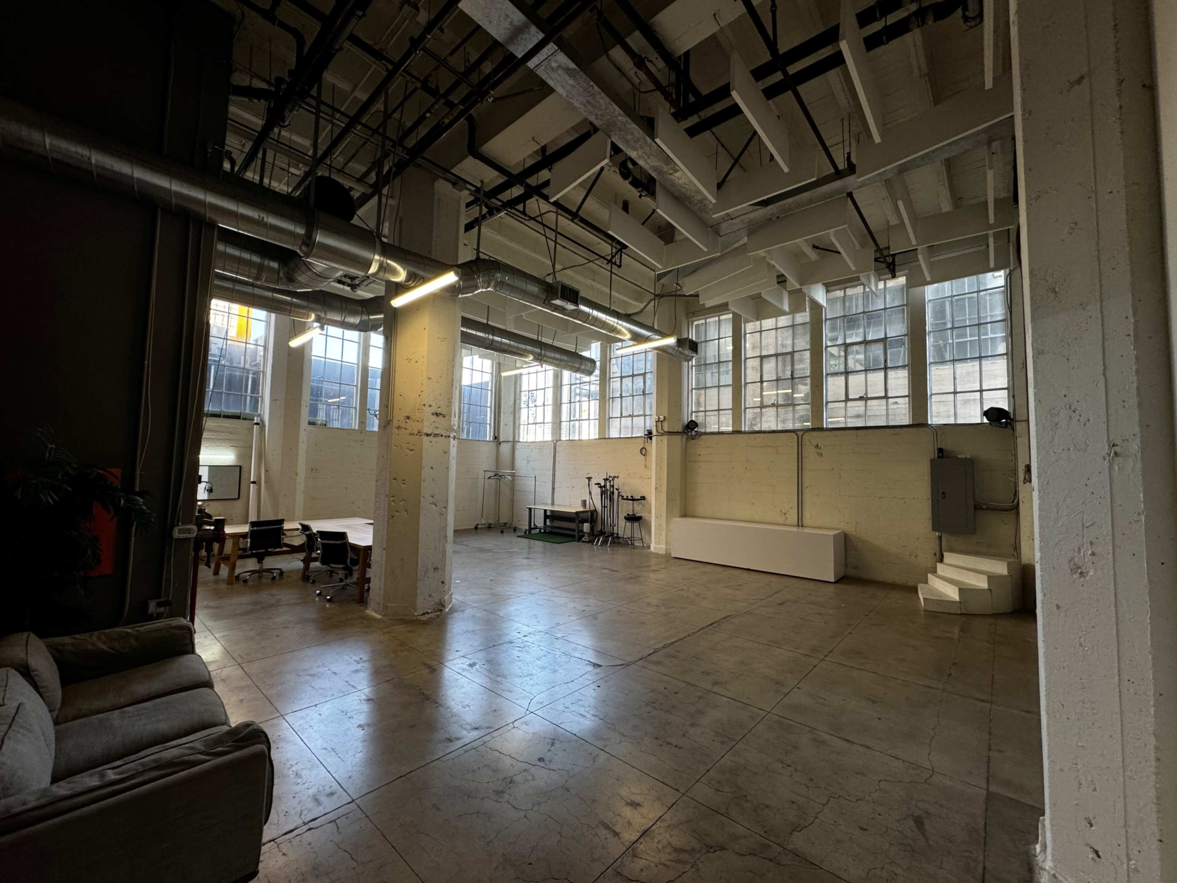 A spacious, industrial-style interior with high ceilings, large windows, and an open layout featuring metal pipes, a seating area, and various workstations.