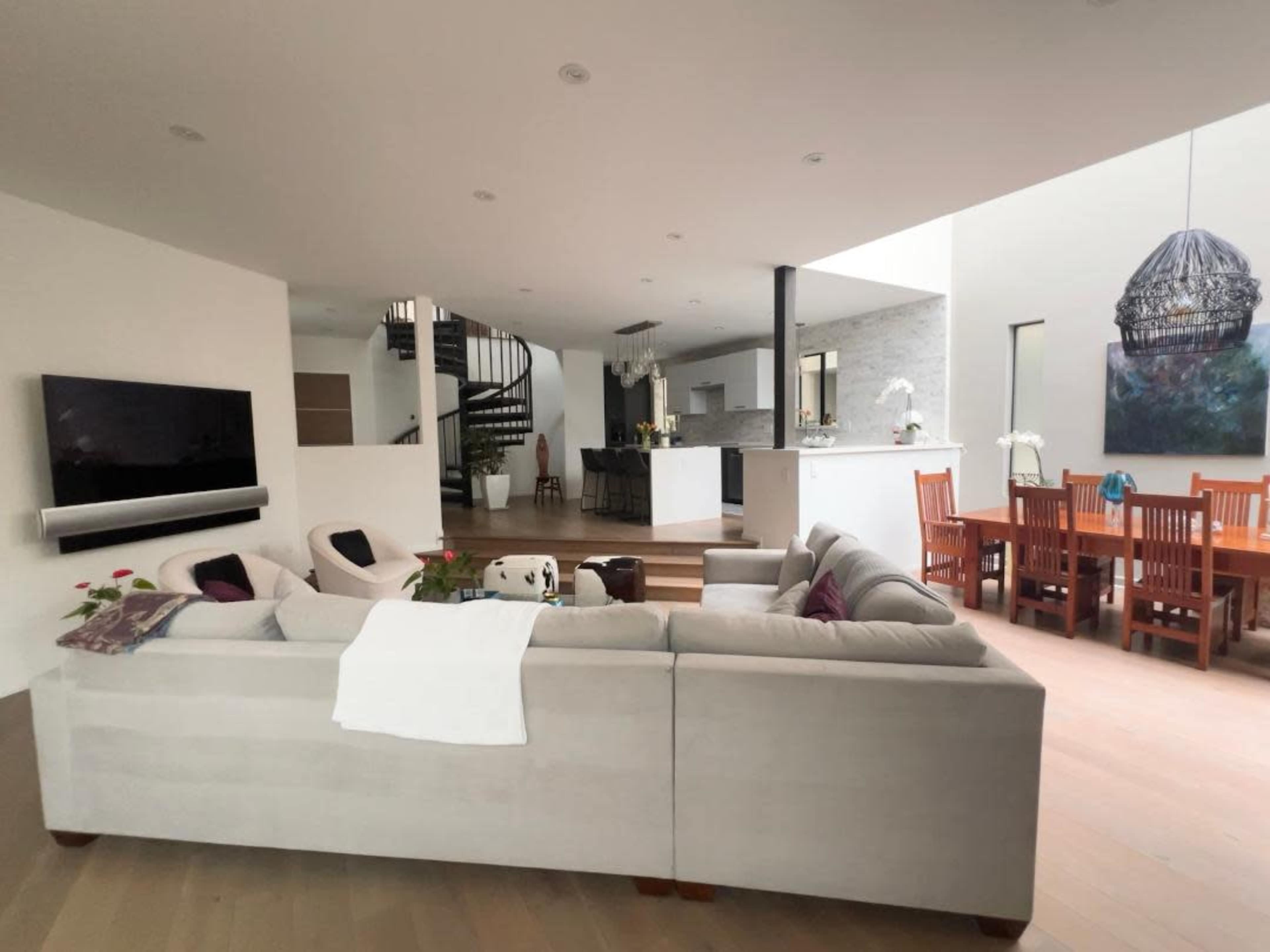 The image shows a modern living room featuring a large sectional couch, a dining area with wooden chairs, and a staircase leading to an upper level.