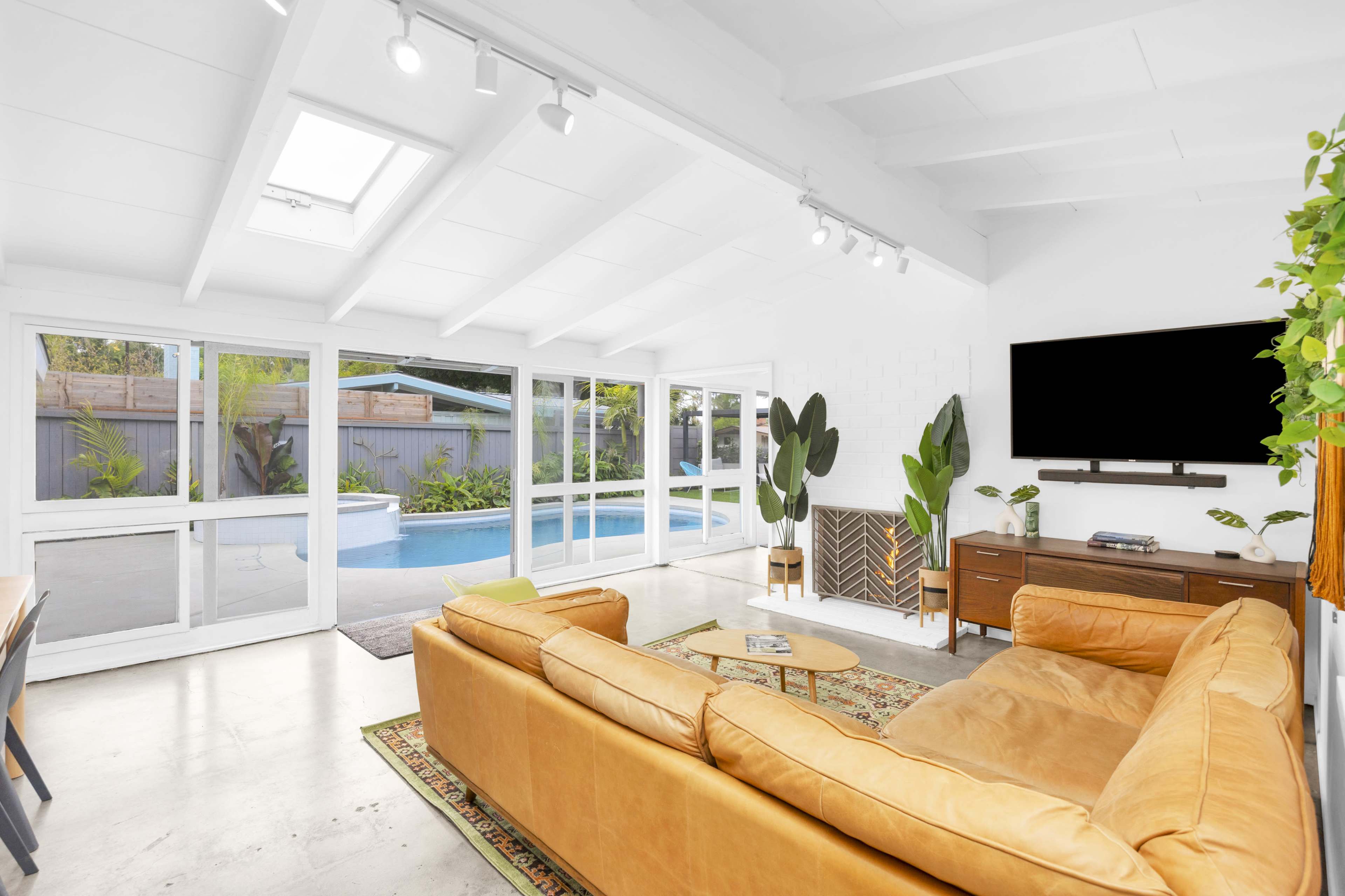 A spacious living area features a large leather sofa, a flat-screen TV mounted on the wall, and sliding glass doors that open to a pool area.