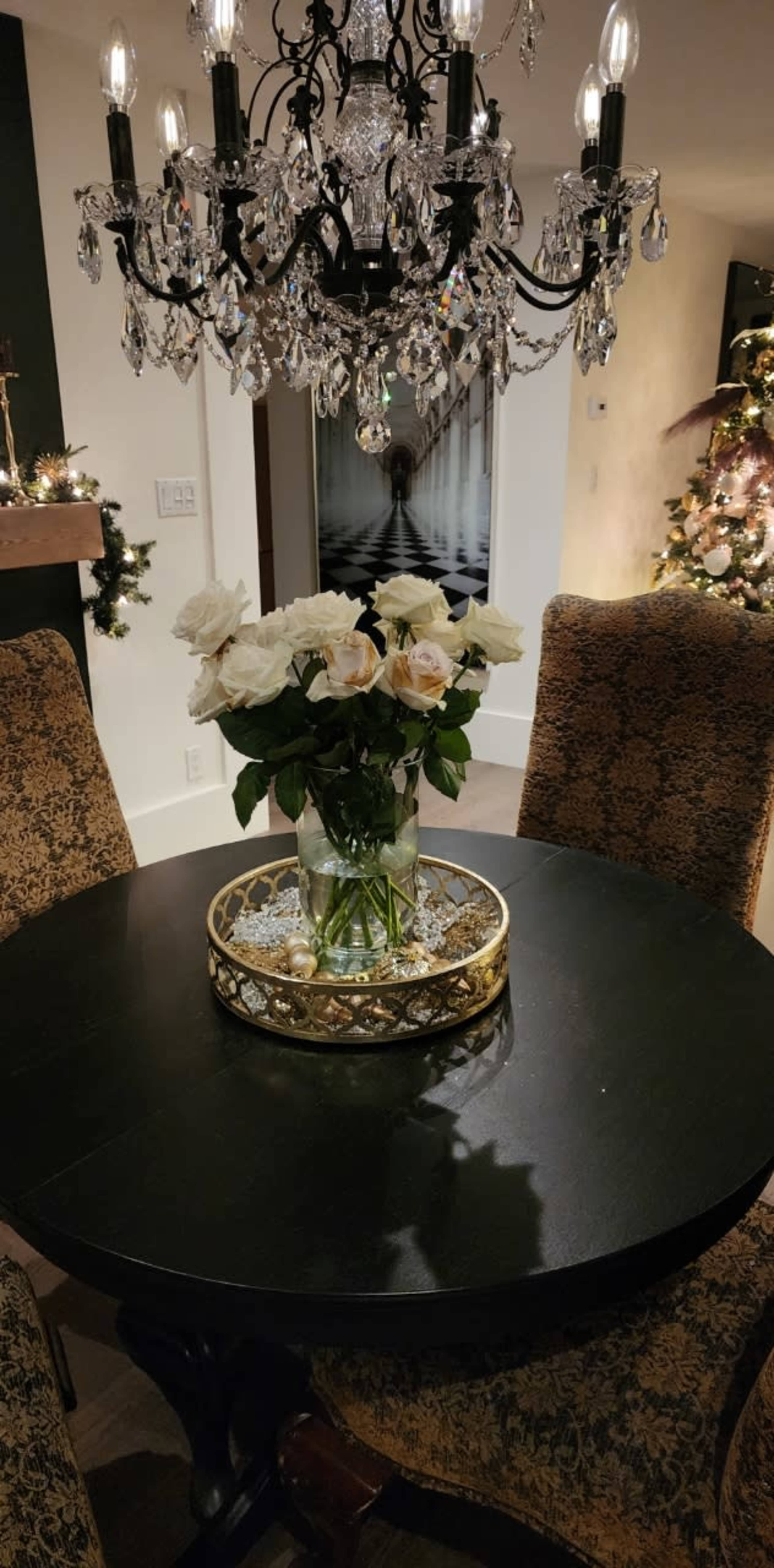 A round black table with a floral arrangement in a glass vase is set under a sparkling chandelier, surrounded by upholstered chairs in a well-lit dining area.