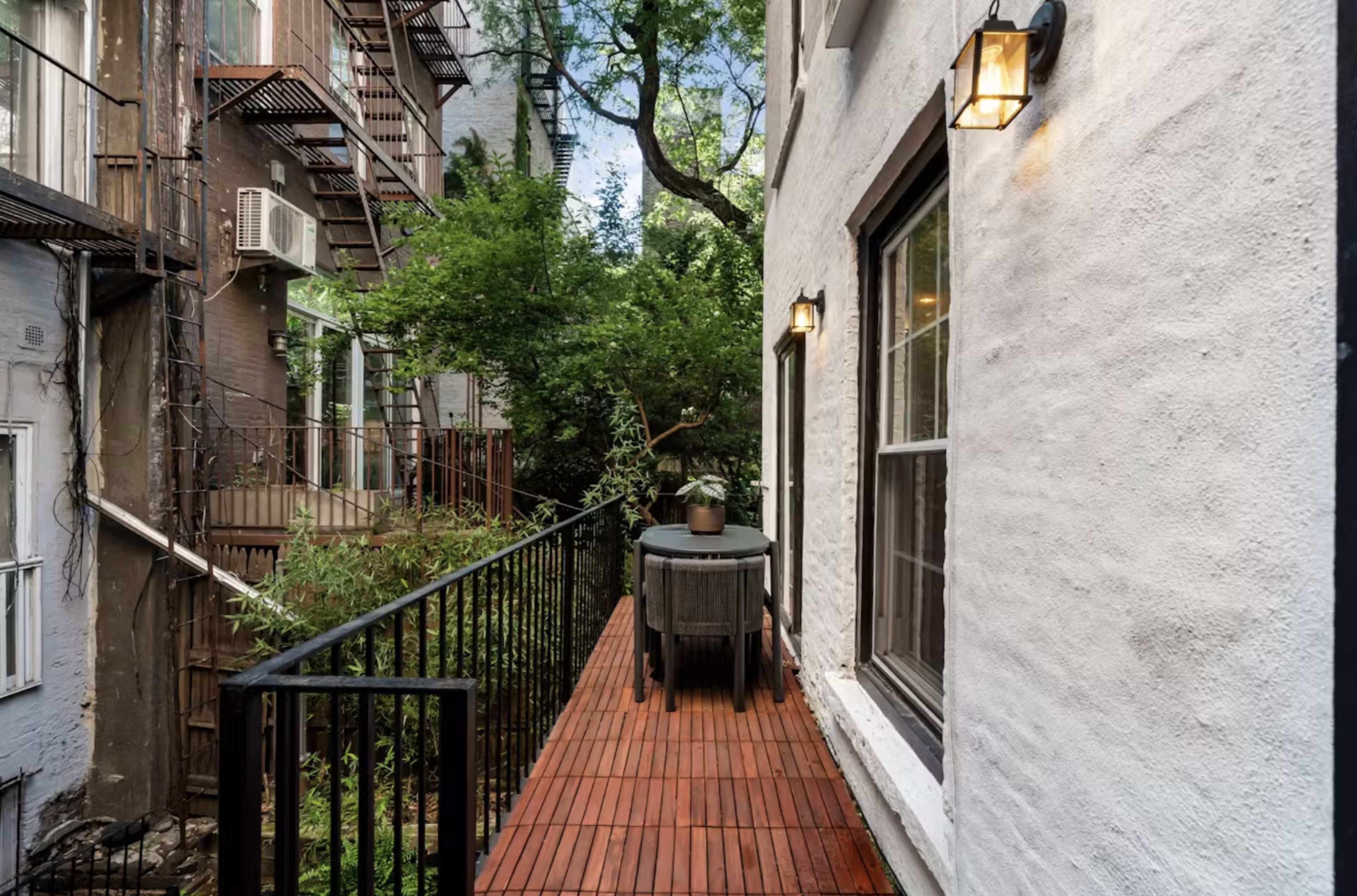 The image shows a narrow wooden walkway lined with black railings, flanked by two well-lit white buildings and greenery.