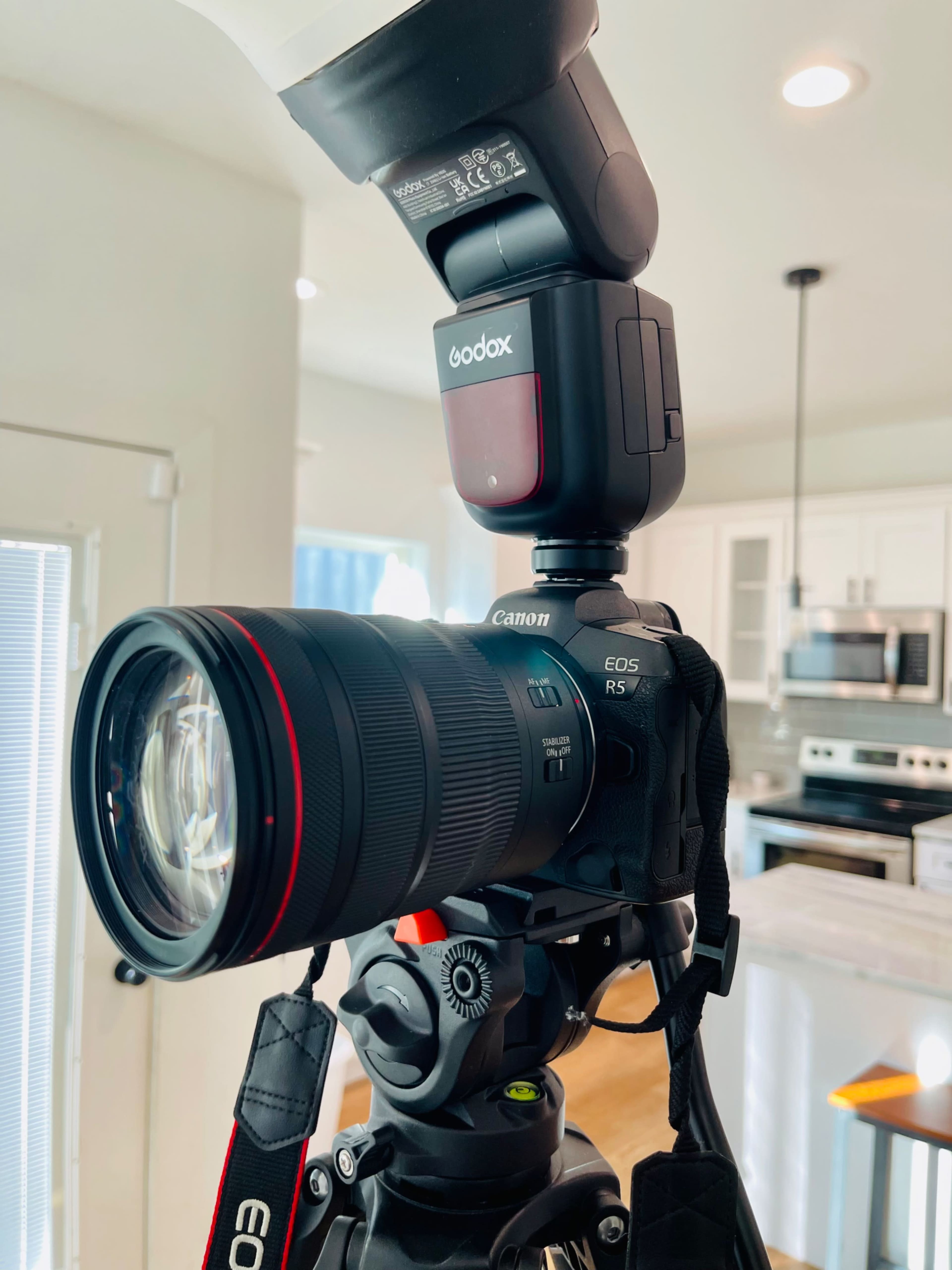 A Canon EOS R5 camera is mounted on a tripod with a Godox flash attached, set up in a kitchen environment.
