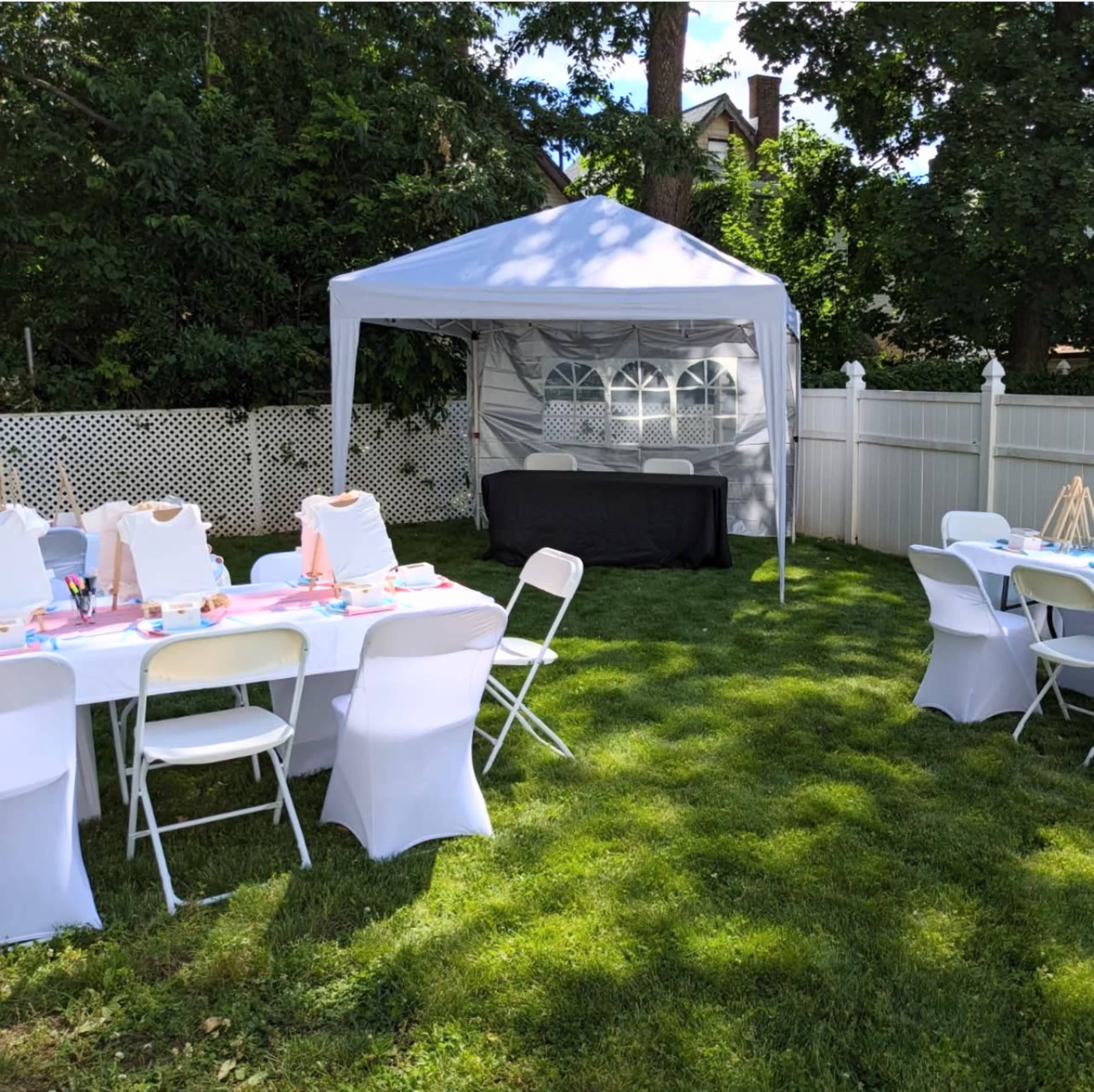 A white canopy covers a black table in a grassy area surrounded by white chairs and a fence.