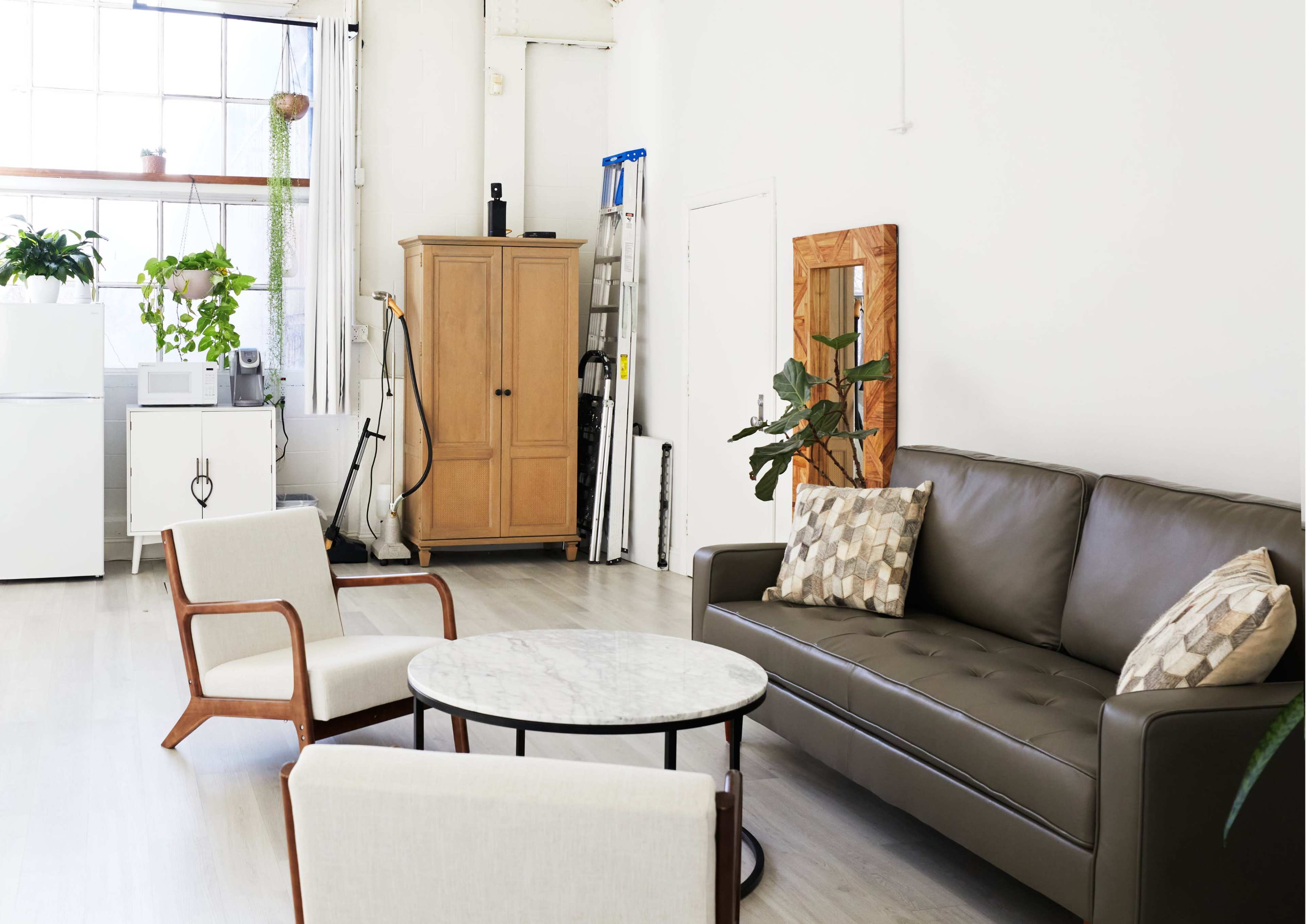 A modern, minimalist living space features a leather sofa, two wooden chairs, a round marble coffee table, and large windows with natural light.