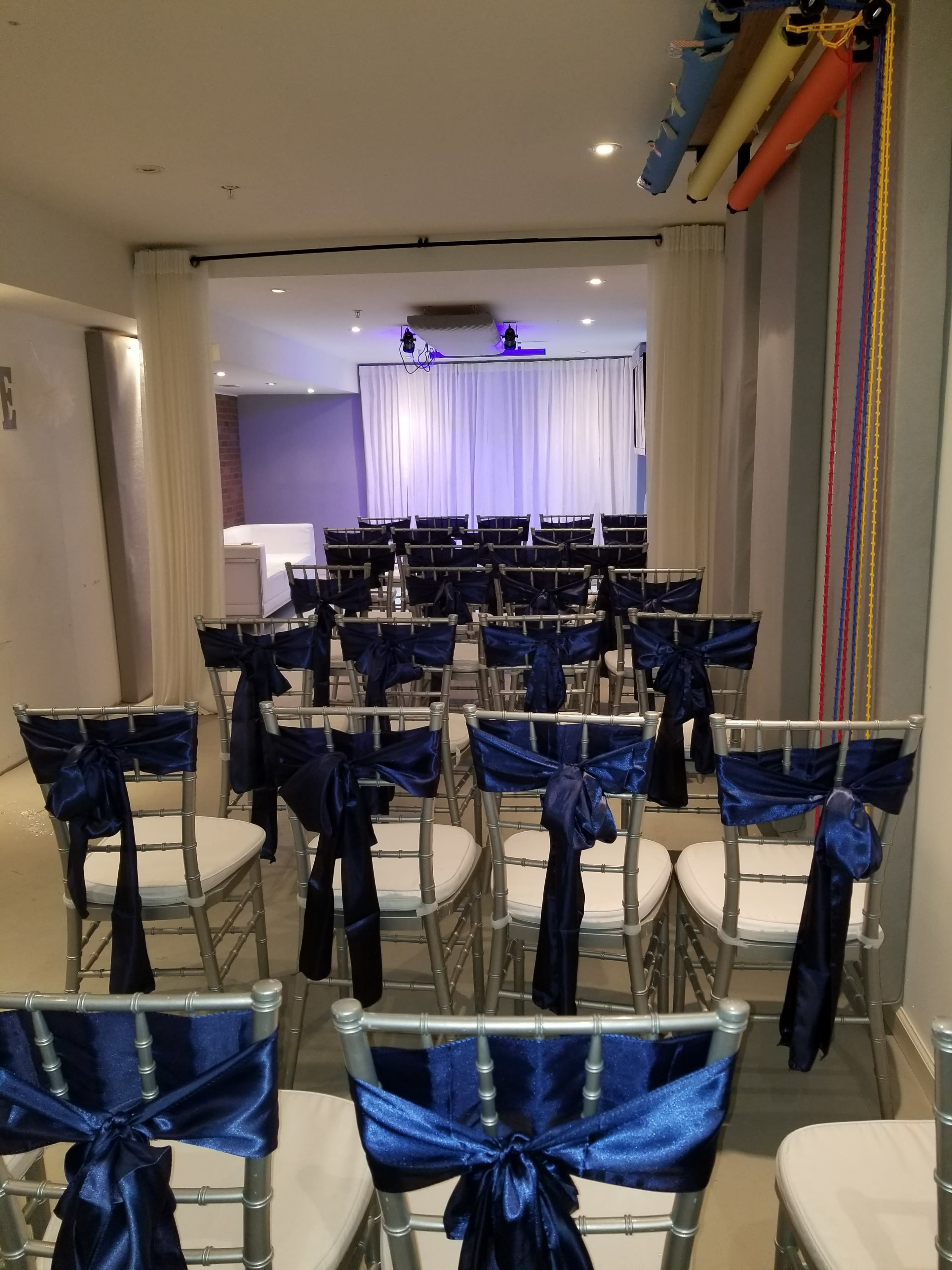 A room set up for an event, featuring rows of chairs with navy blue sashes facing a stage with white curtains in the background.