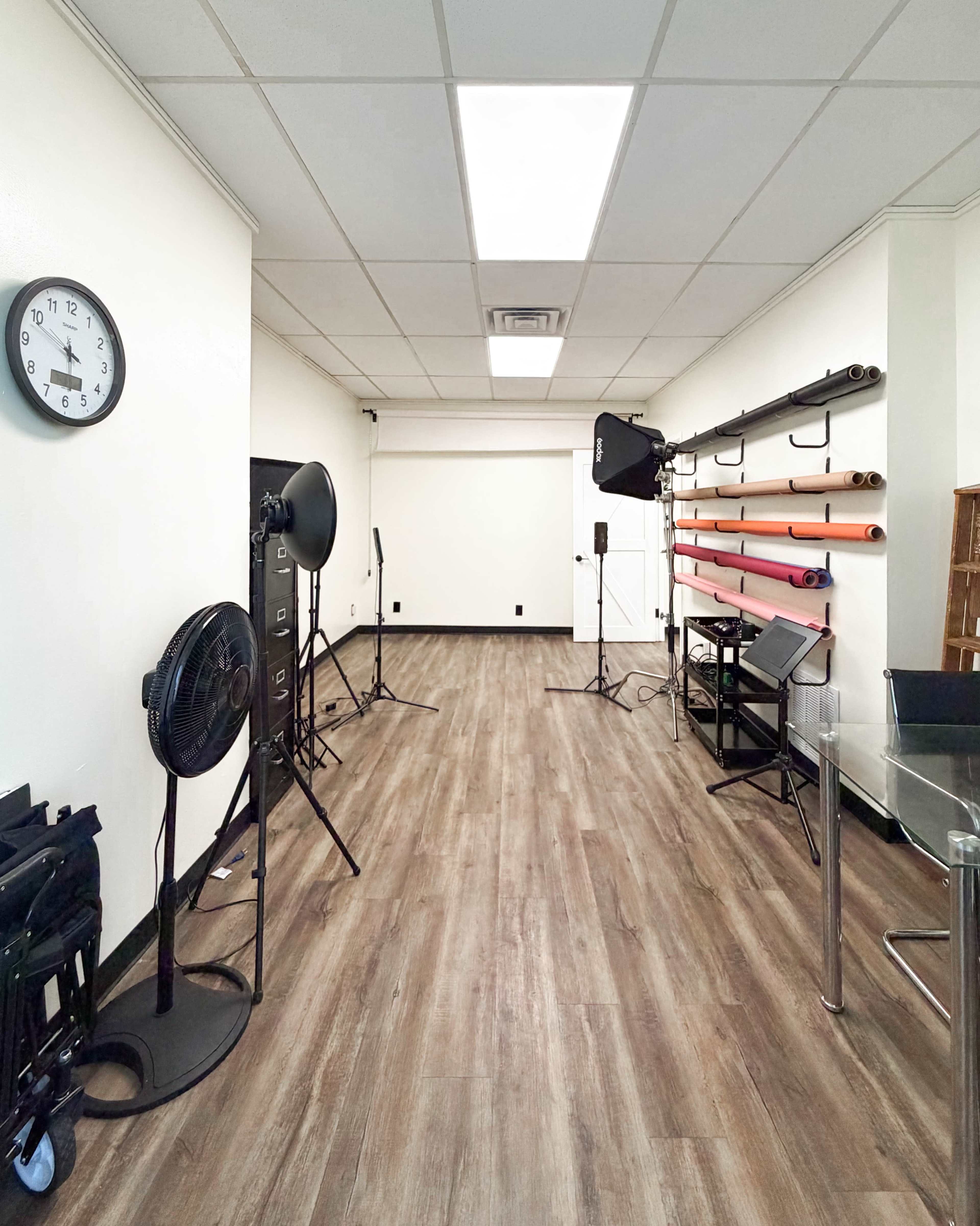 The image shows a well-lit studio space with photography equipment, including softboxes, background rolls, and a clock on the wall.