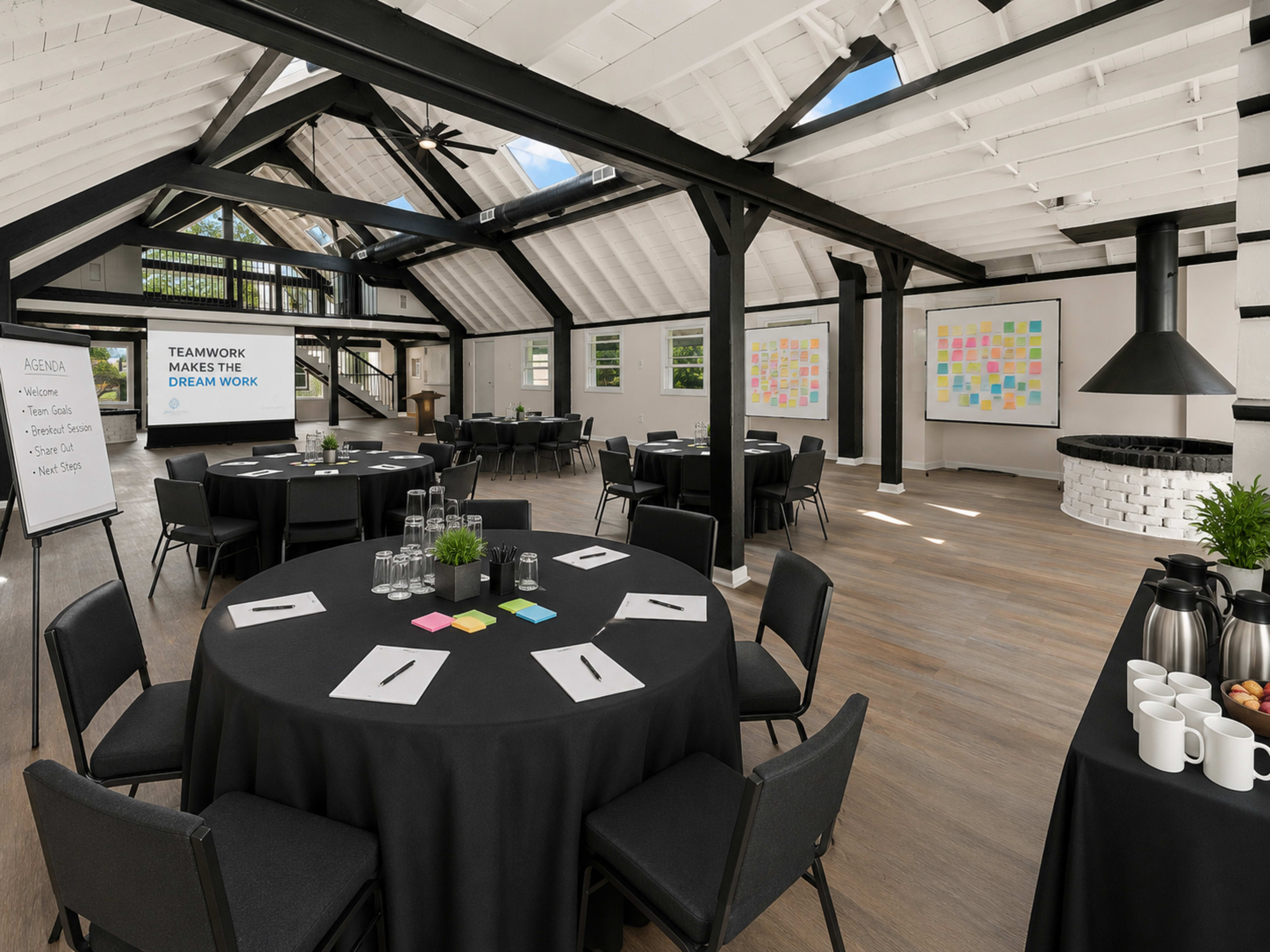 The image shows a spacious conference room with round tables covered in black tablecloths, equipped with chairs, notepads, and colorful sticky notes, alongside a presentation area featuring two large whiteboards.