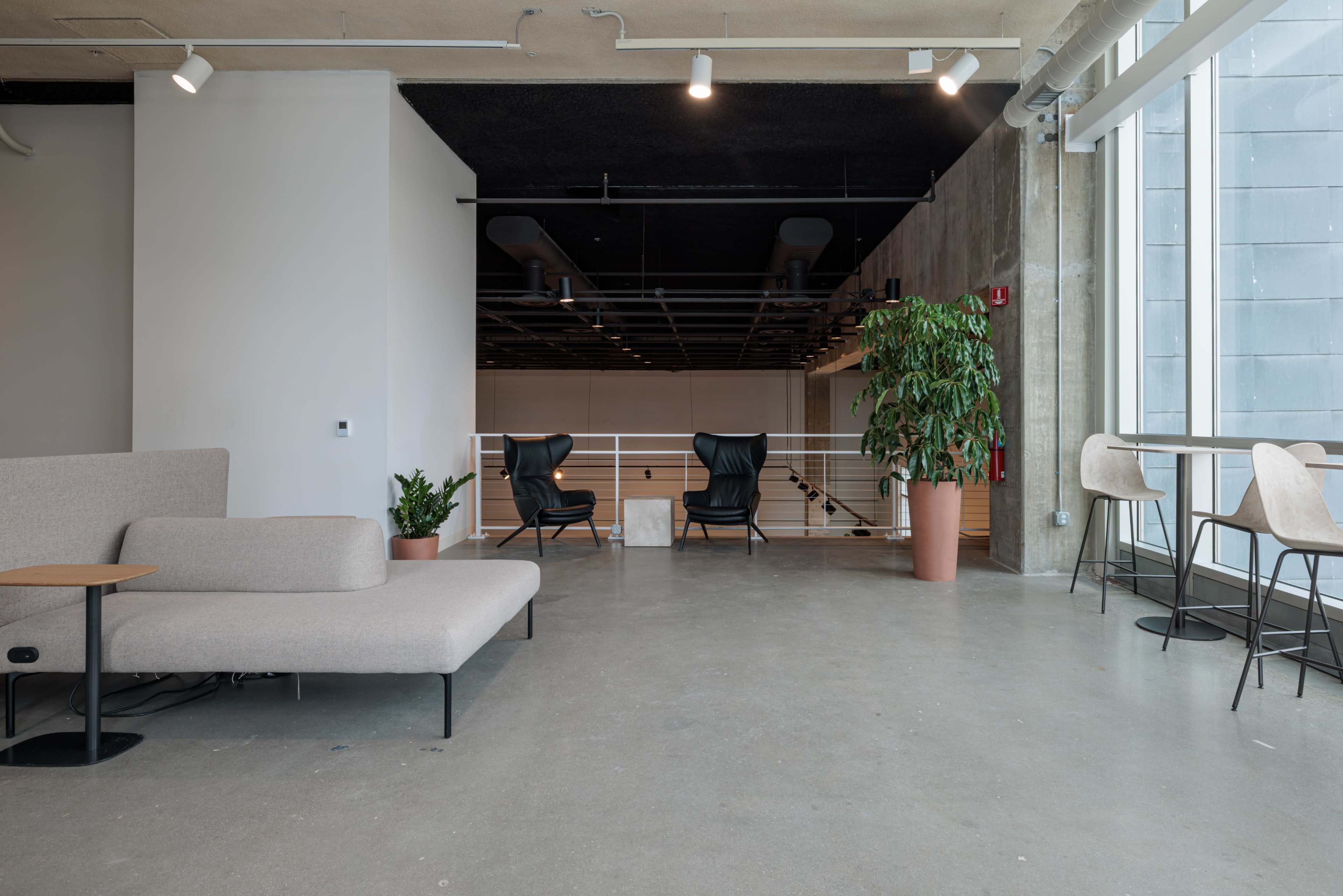 The scene features a modern, minimalist interior with a gray sofa, two armchairs, a potted plant, and a few barstools near a large window.