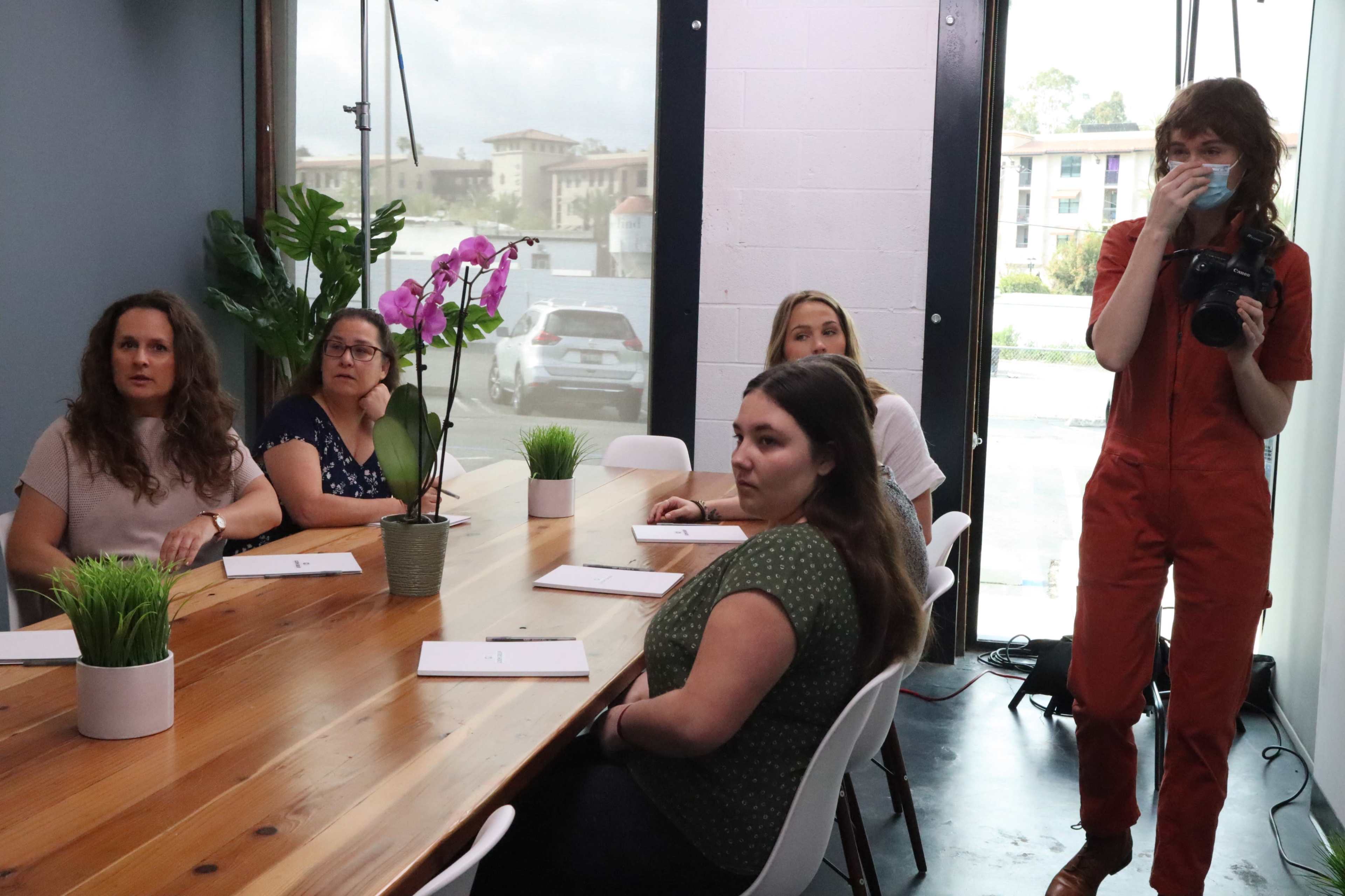 A group of four women sits at a wooden table with potted plants, while a woman in an orange jumpsuit stands nearby holding a camera.