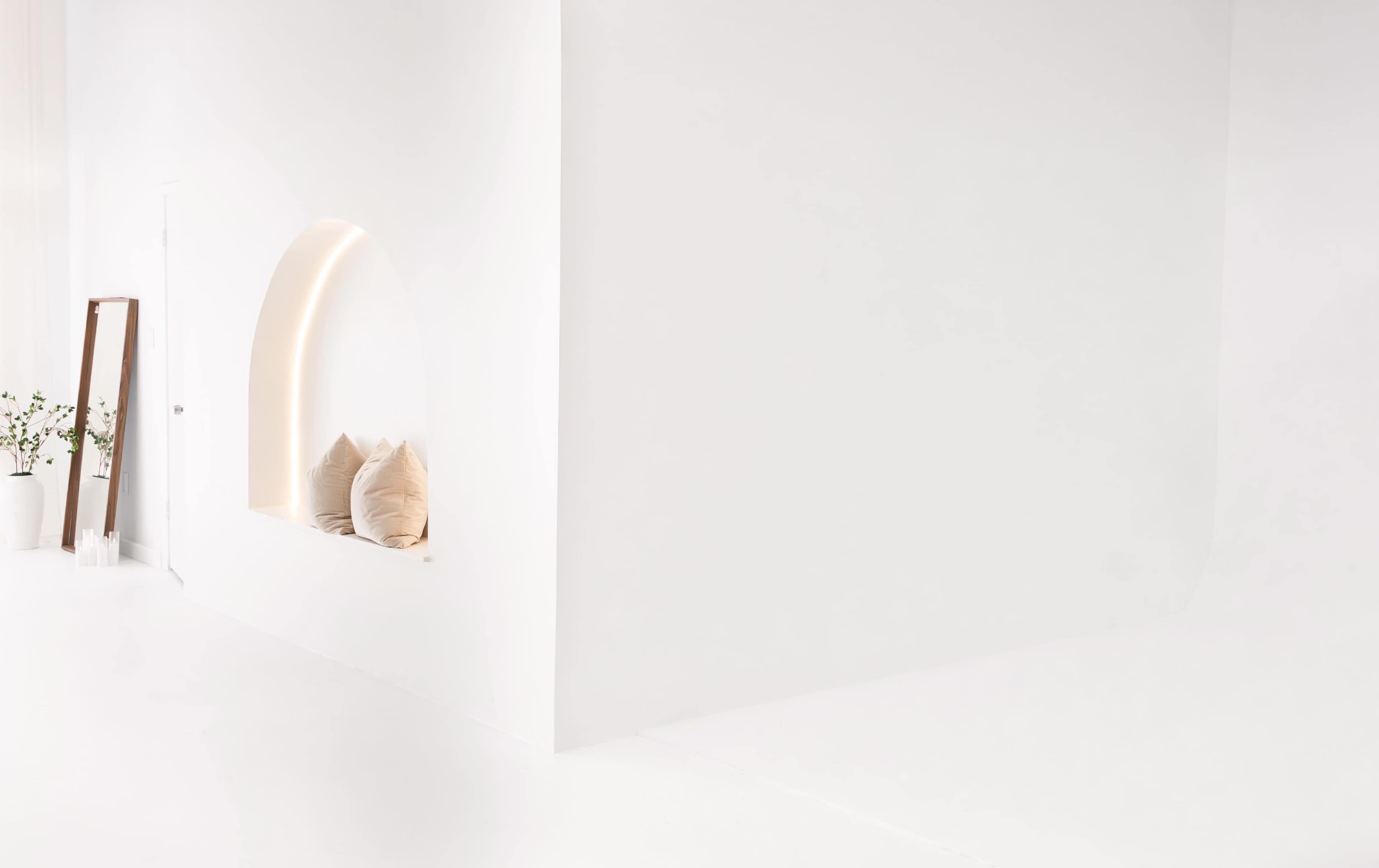The image features a minimalist interior with a white wall, an arched nook containing two pillows, and a tall mirror beside a potted plant.