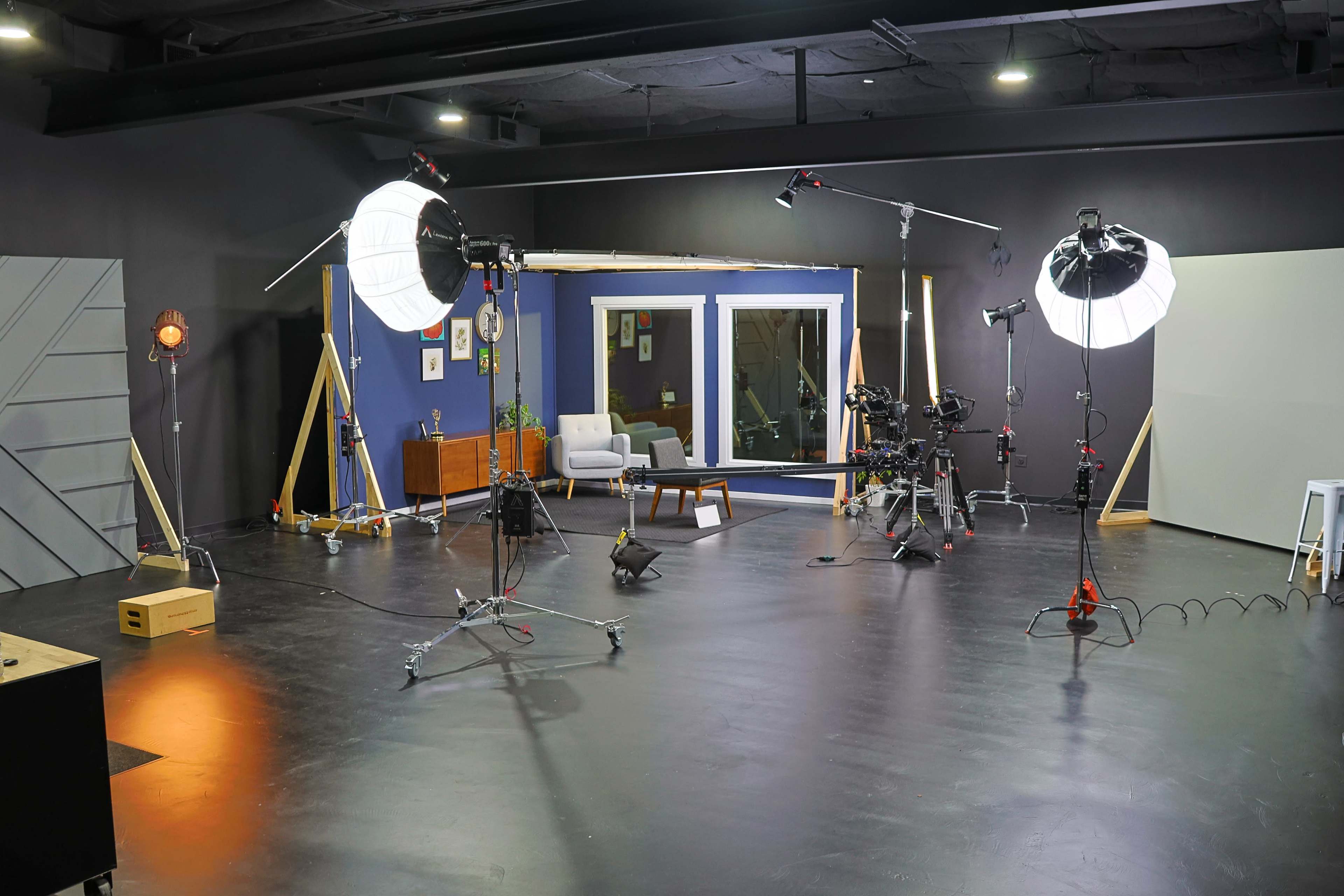 A photography studio is set up with various lighting equipment, cameras, and a staged area featuring a chair and a window.