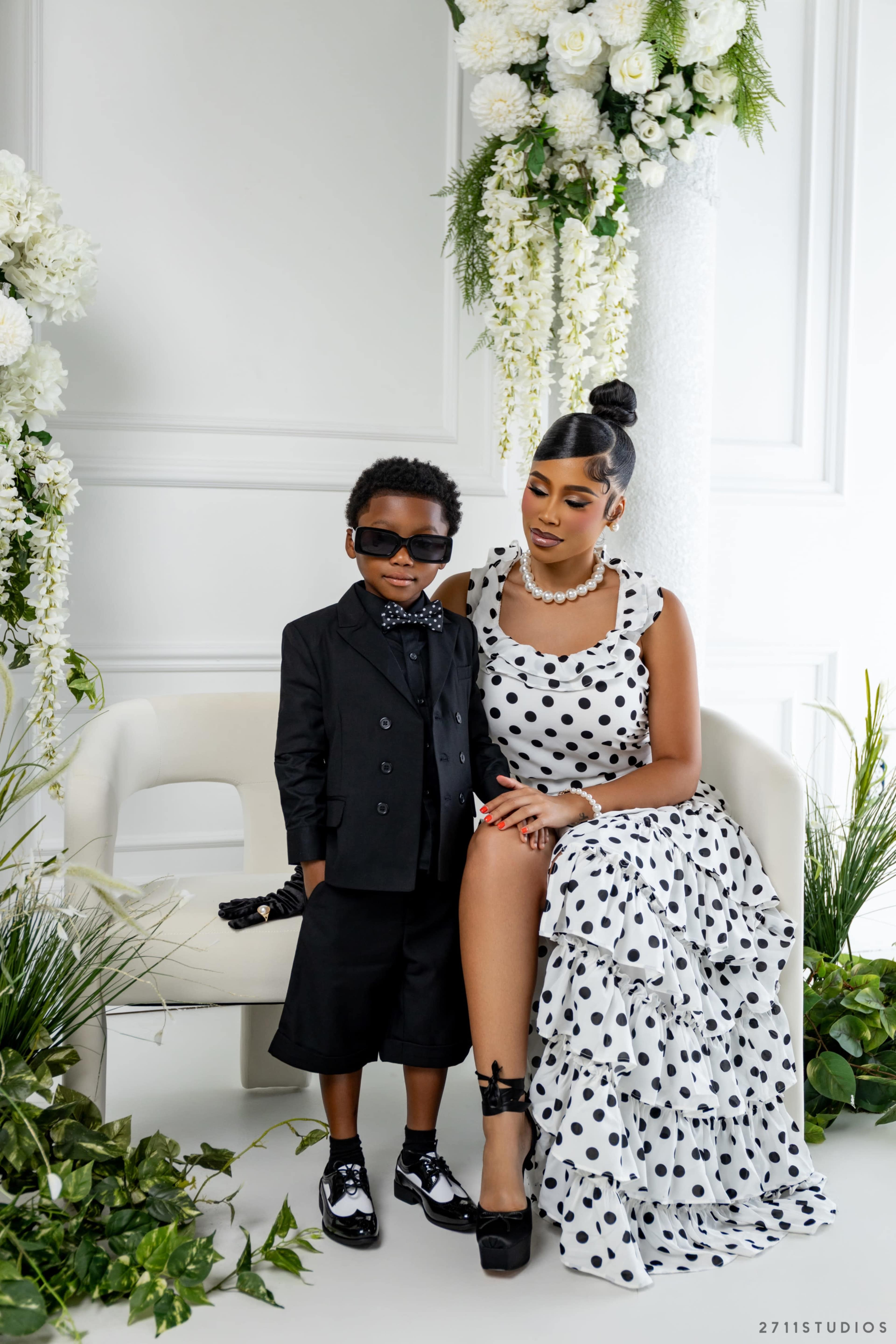 A boy in sunglasses stands next to a woman dressed in a polka dot gown, both posing in a floral-themed setting.