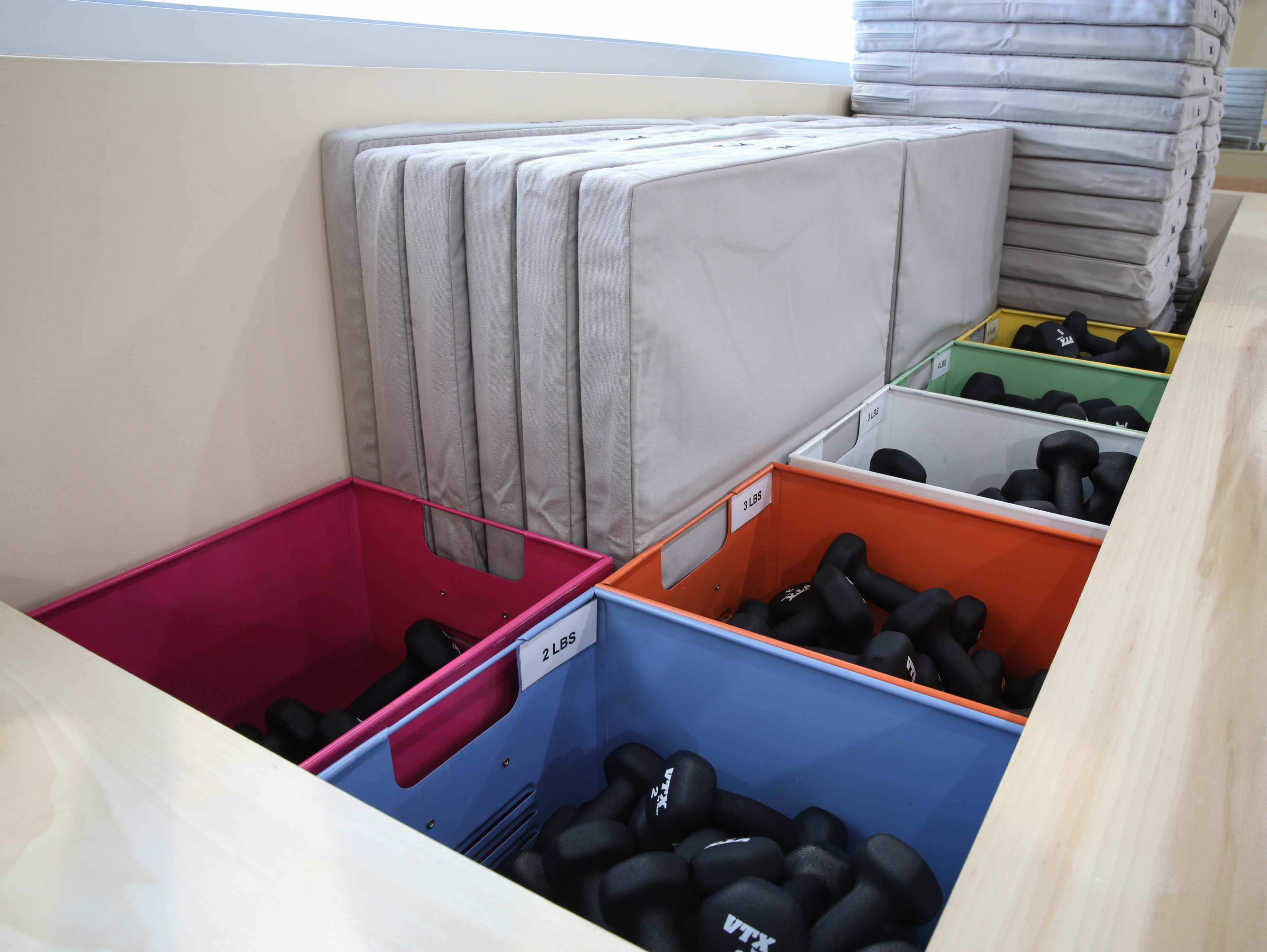 A collection of colorful bins filled with black dumbbells of various weights, along with stacked cushions above the bins.