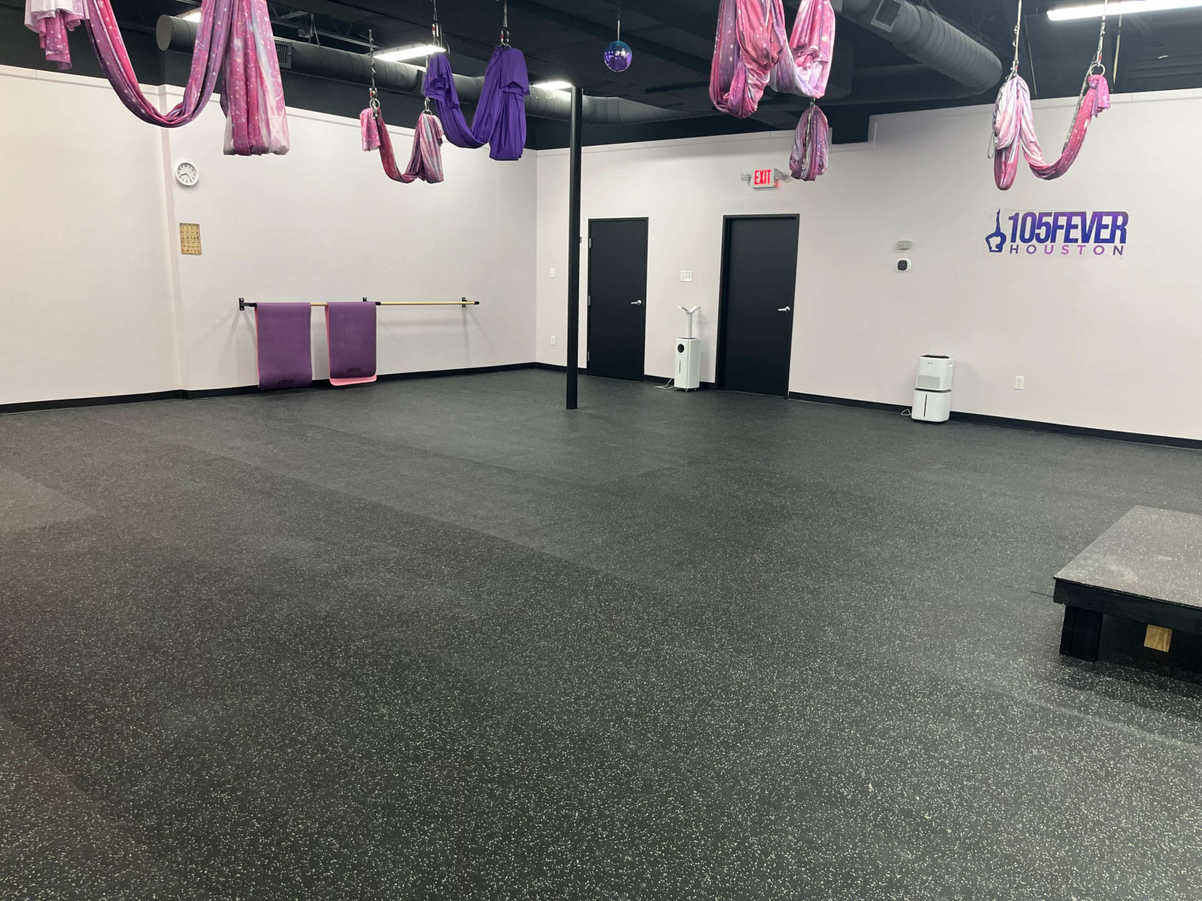 A spacious, empty fitness studio with dark flooring, pink aerial hammocks hanging from the ceiling, and a counter with equipment against the wall.