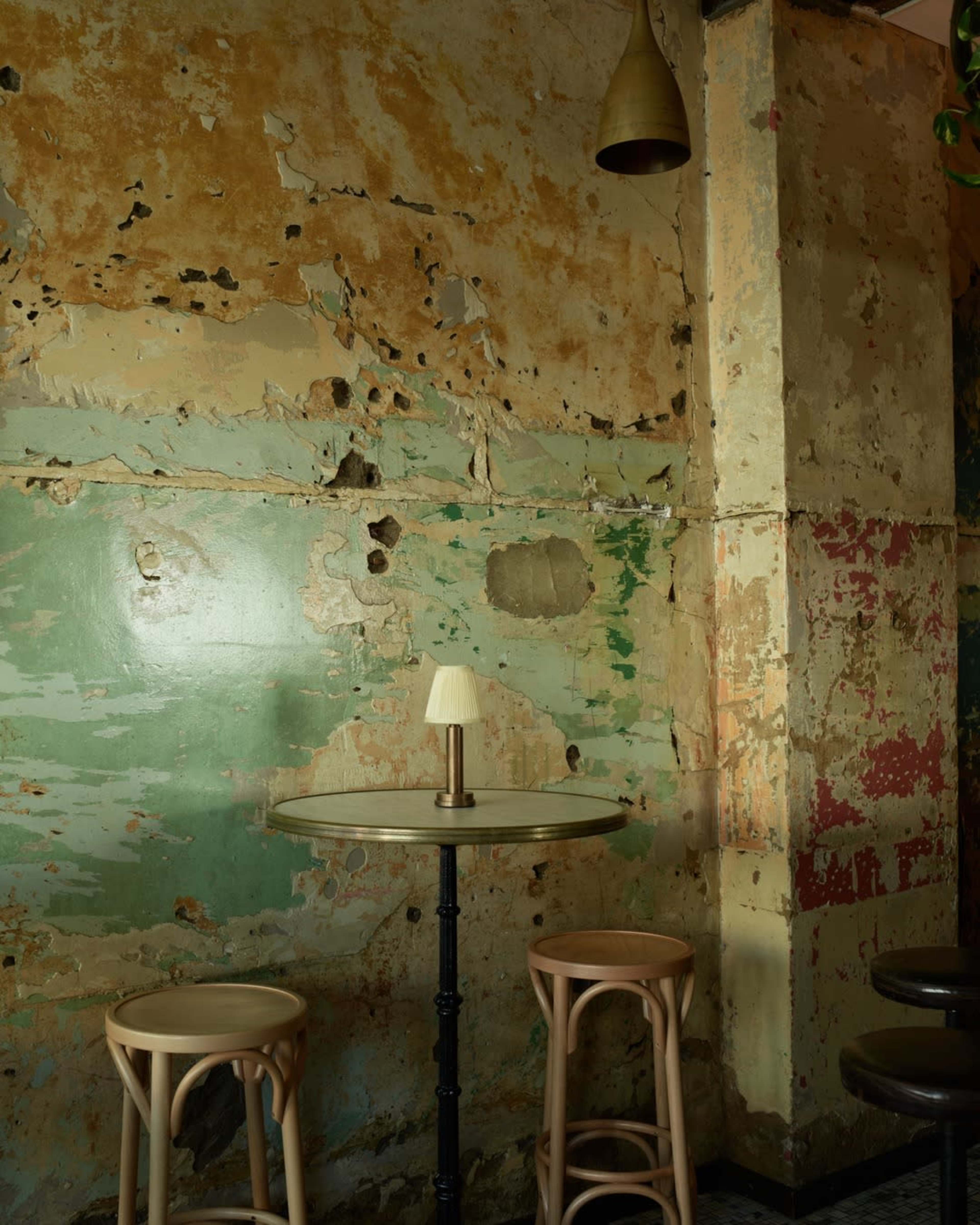 A small round table with a lamp sits against a textured, peeling wall painted in green and beige.