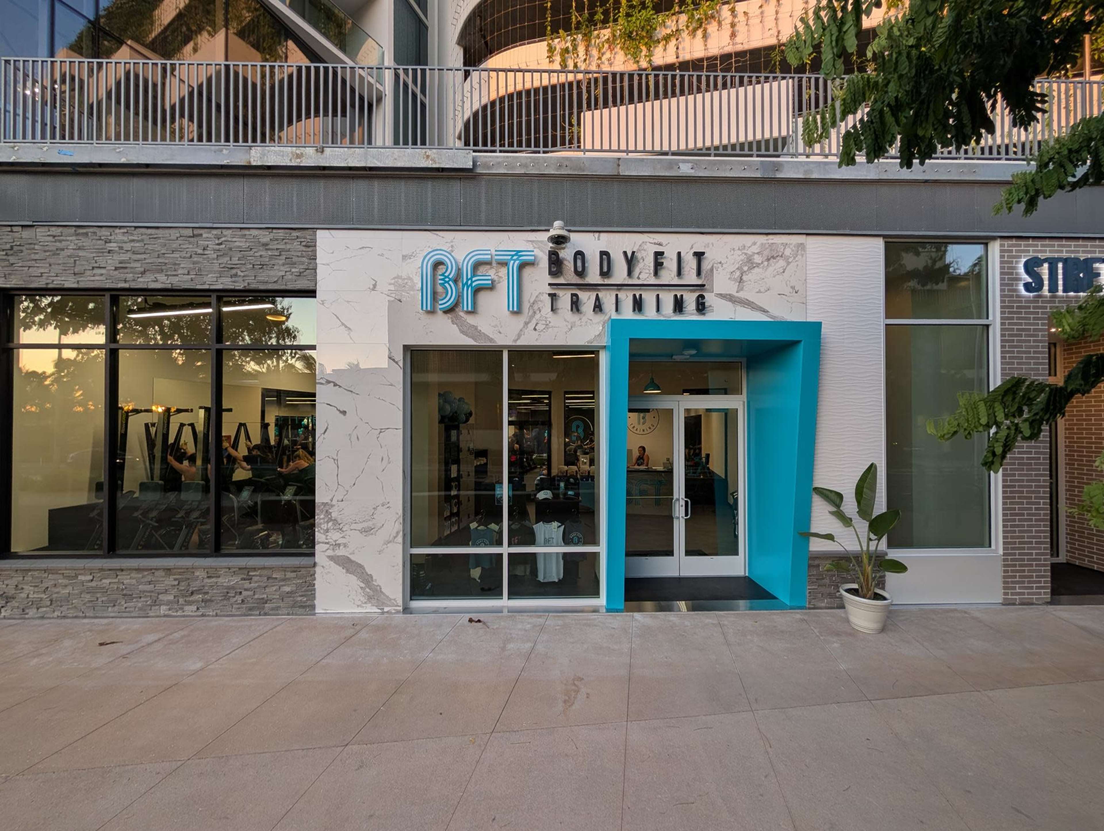 The image shows the exterior of a fitness training studio called BFT Bodyfit Training, featuring large glass windows and a bright blue entrance.