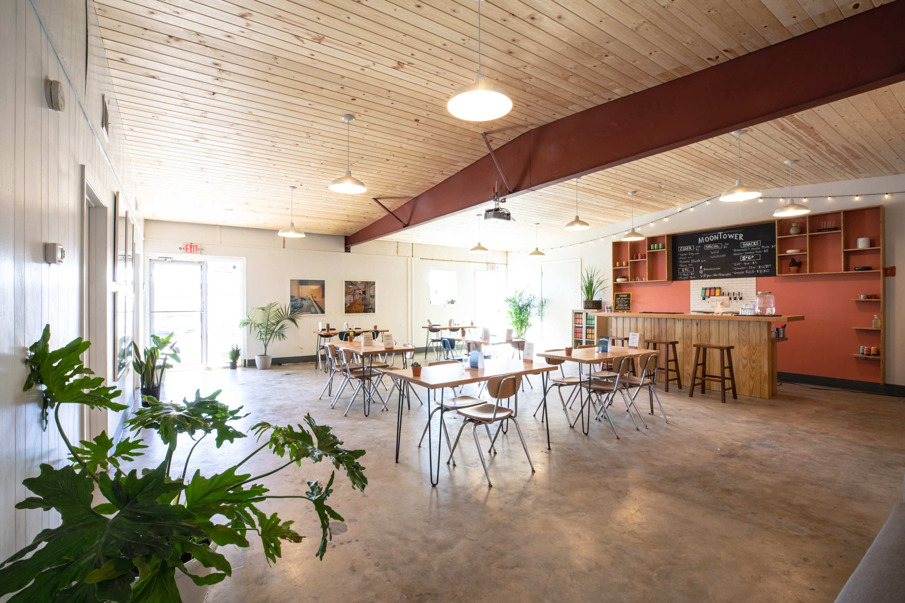 The image shows a spacious interior of a café with wooden ceilings, tables arranged for dining, and a counter featuring shelves of decorative items.