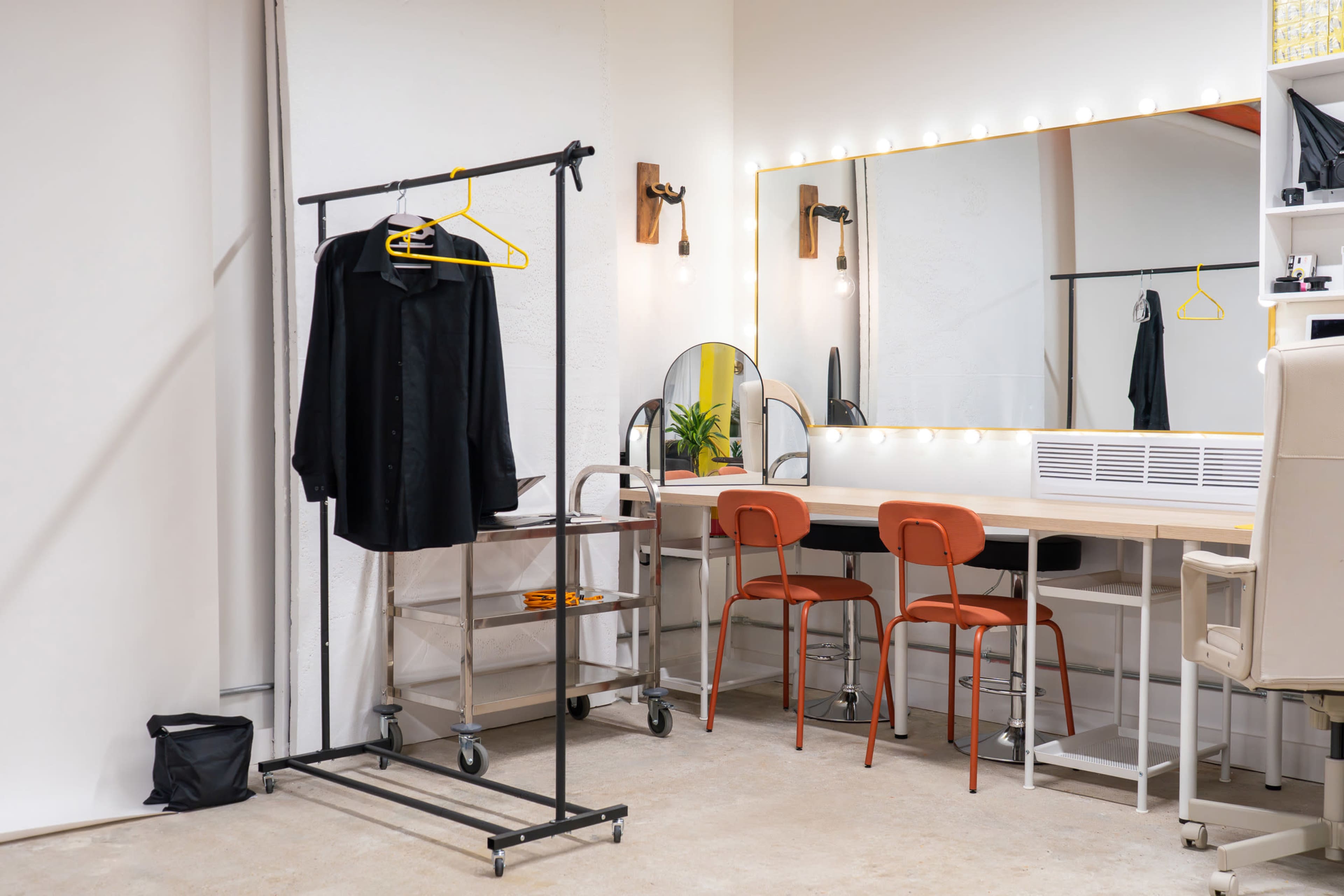 The image shows a dressing area with a black shirt on a clothing rack, a large mirror surrounded by lights, and two orange chairs at a vanity table.