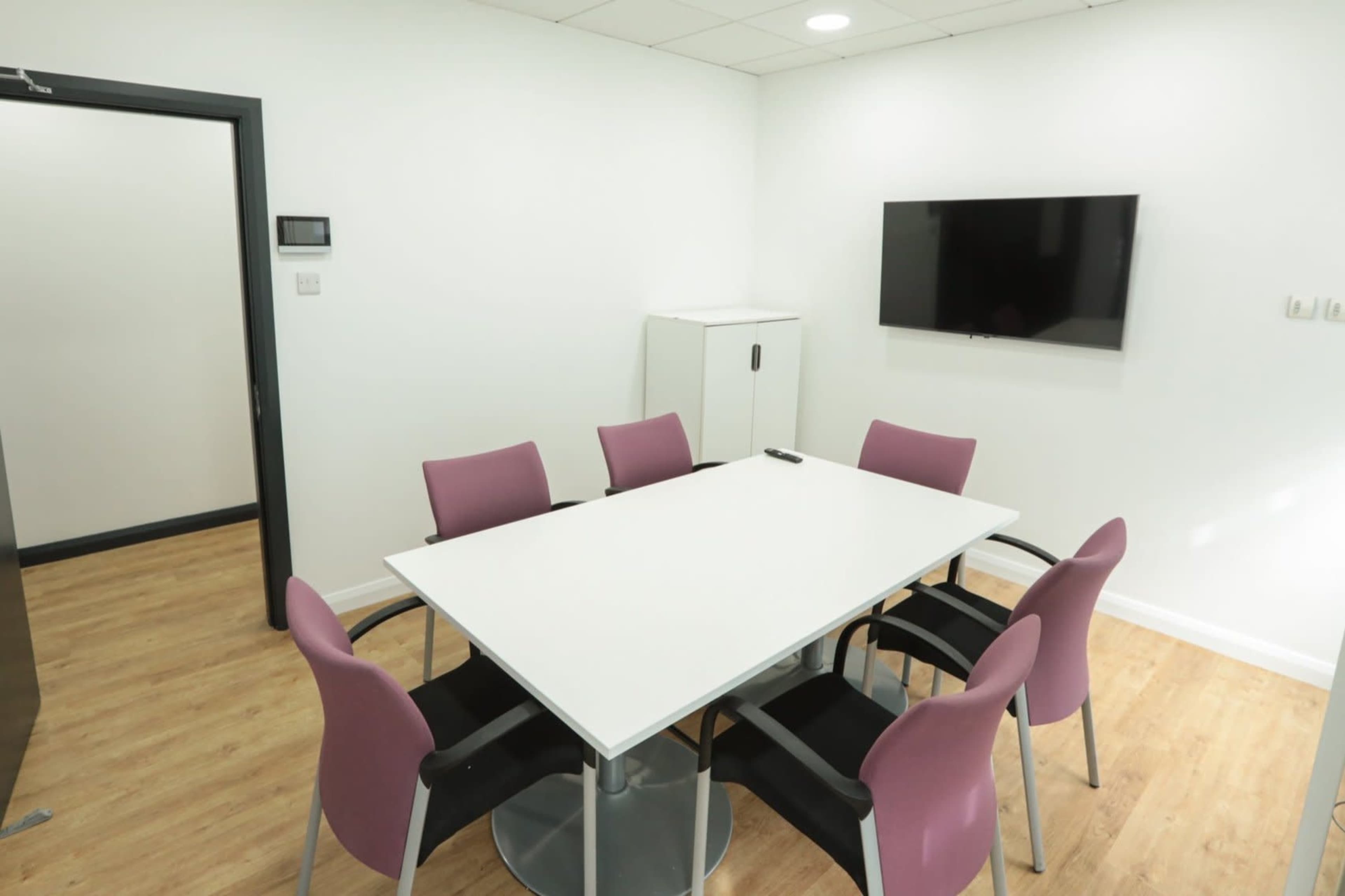A modern meeting room features a rectangular white table surrounded by six chairs, with a wall-mounted television and a cabinet in the background.