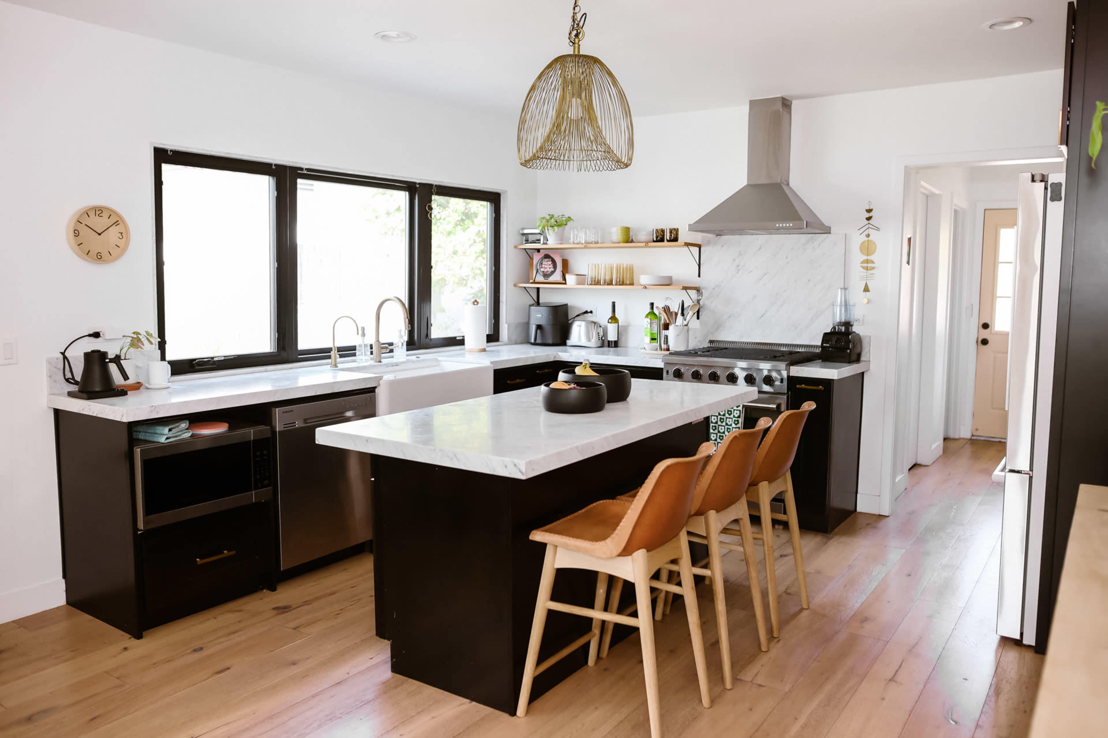 A modern kitchen features a white marble countertop, black cabinetry, and wooden bar stools, with large windows allowing natural light to fill the space.