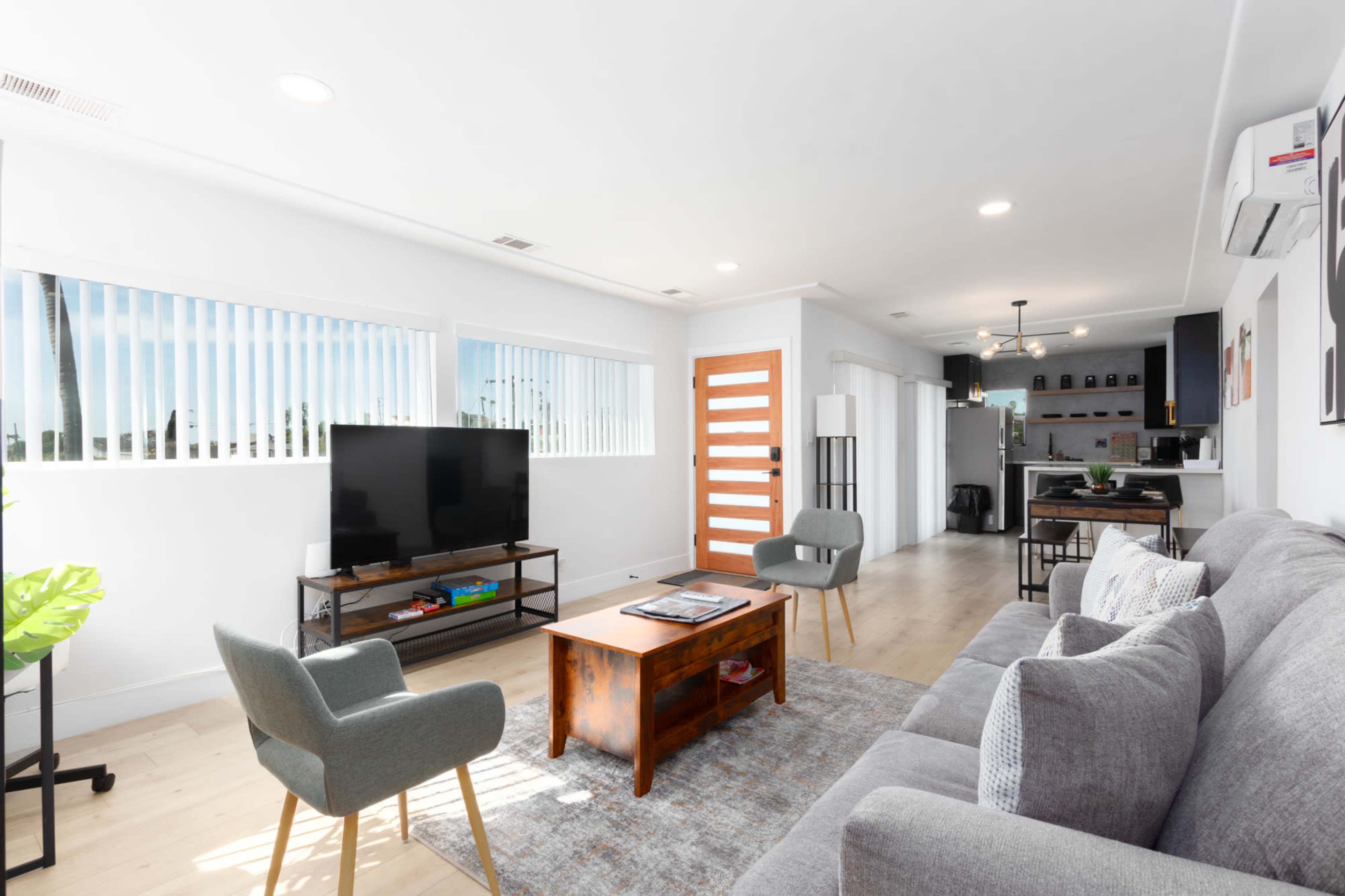 The image shows a modern living room with a gray sofa, a wooden coffee table, a television on a stand, and large windows allowing natural light to enter.