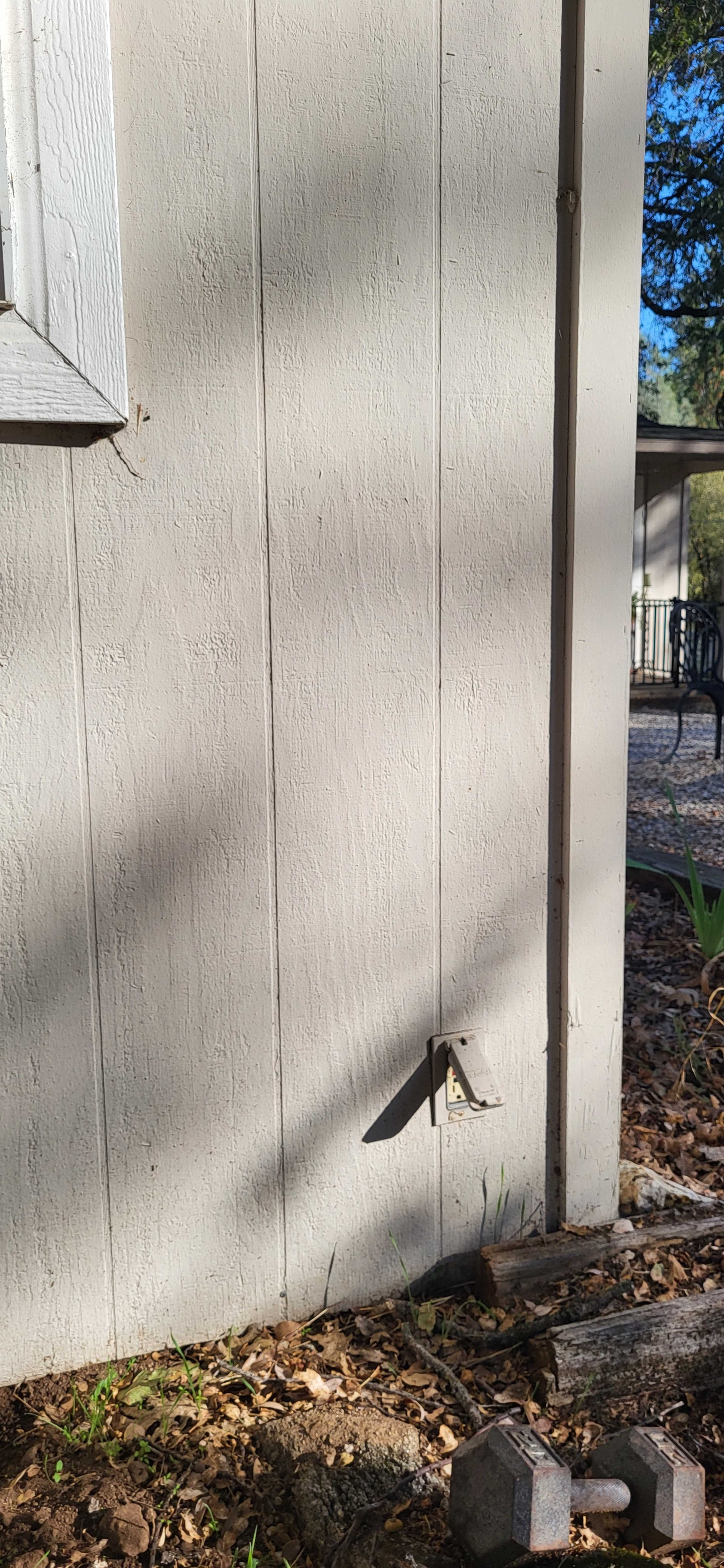 The image shows the exterior of a light-colored wooden shed wall with a small metal bracket and shadow cast on it, along with some leaves and a dumbbell on the ground nearby.