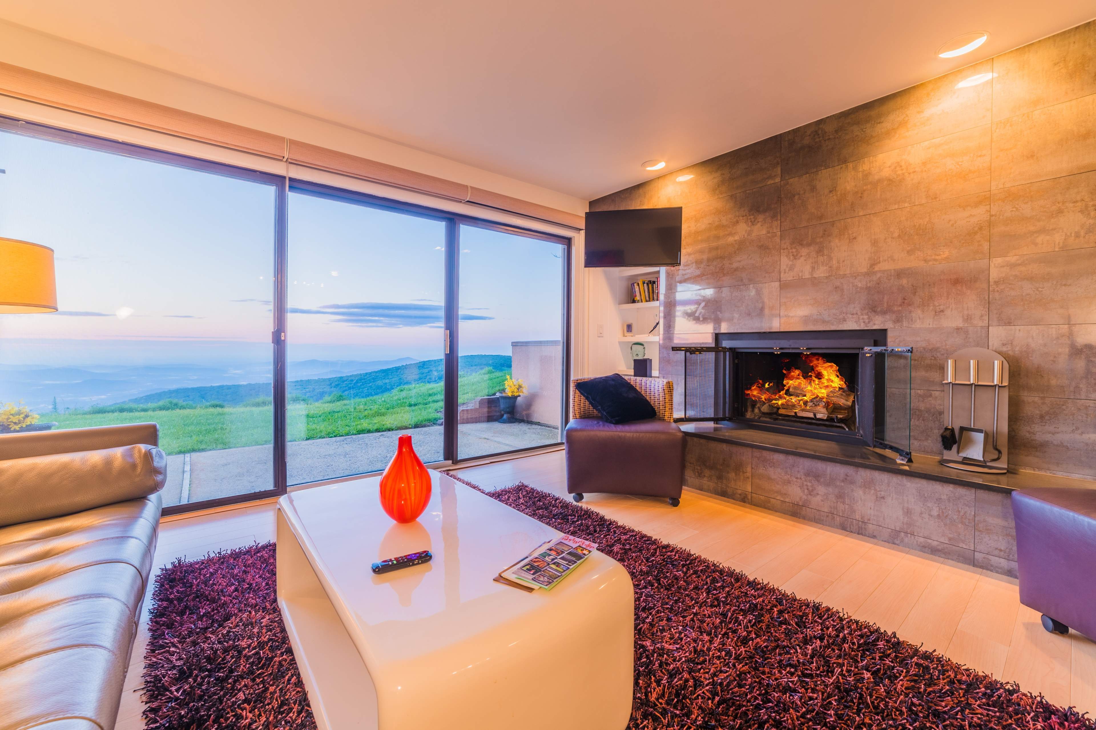 A modern living room features a large window overlooking a scenic view, a fireplace, a coffee table, and contemporary furniture.