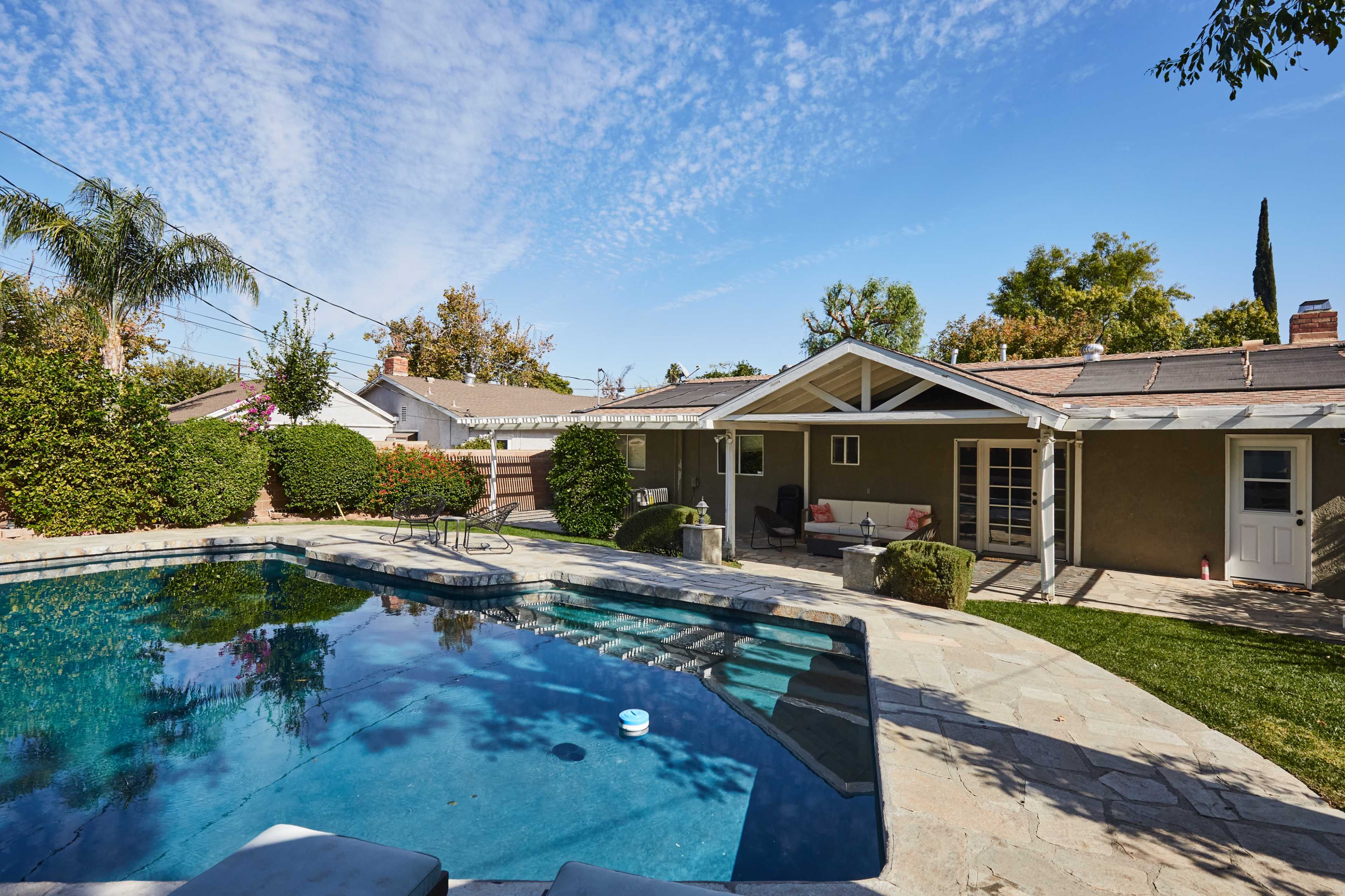 A backyard features a swimming pool surrounded by stone decking and greenery, with a house visible in the background.