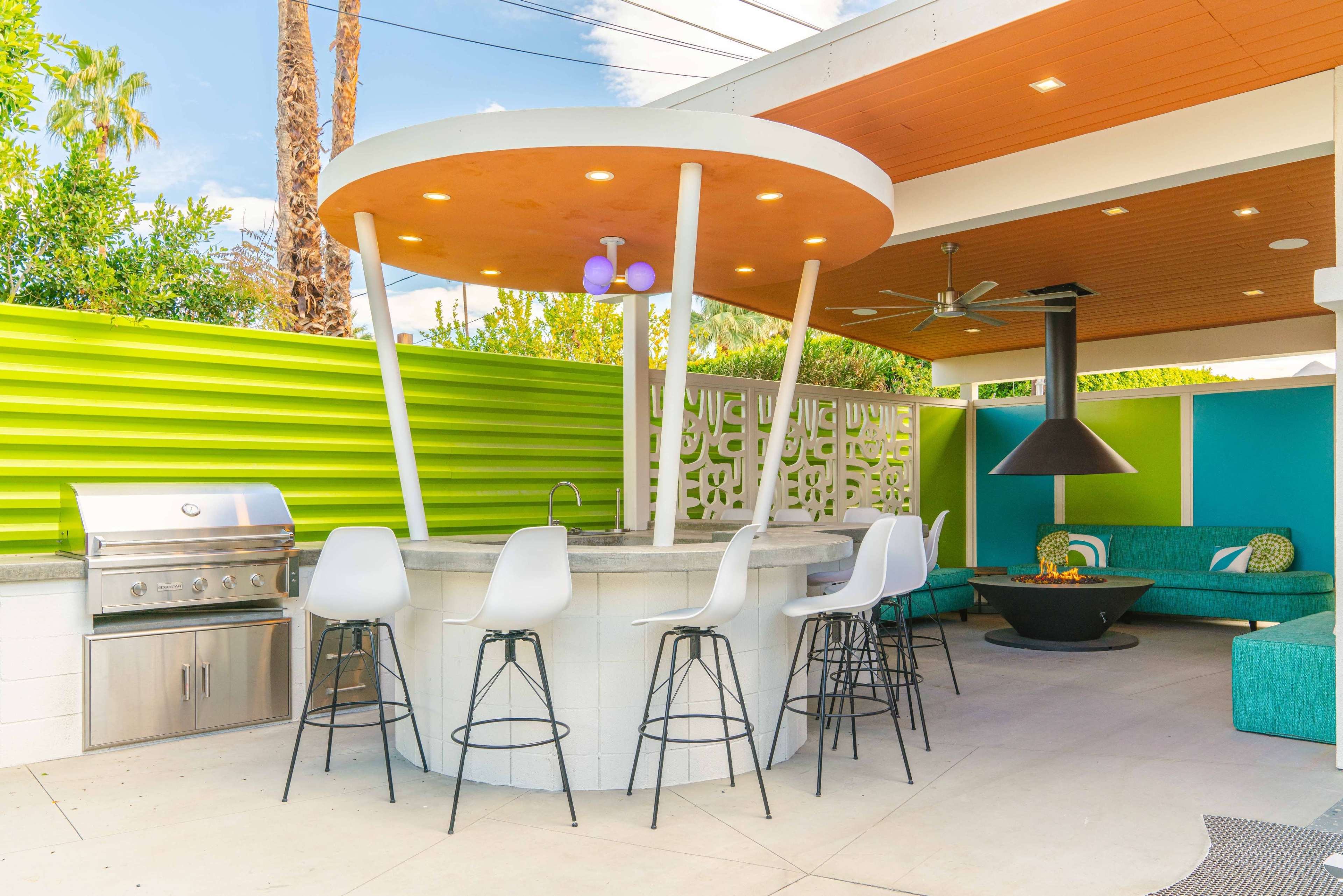 The image shows a modern outdoor space featuring a circular bar with stools, a grill, and a lounge area with a fire pit under a shaded roof.